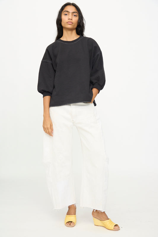 A person stands against a white background wearing the Rachel Comey Fond Sweatshirt in Charcoal, which features an oversized fit and raw edge hemline, paired with loose white pants and yellow open-toe shoes, with one hand in their pocket.