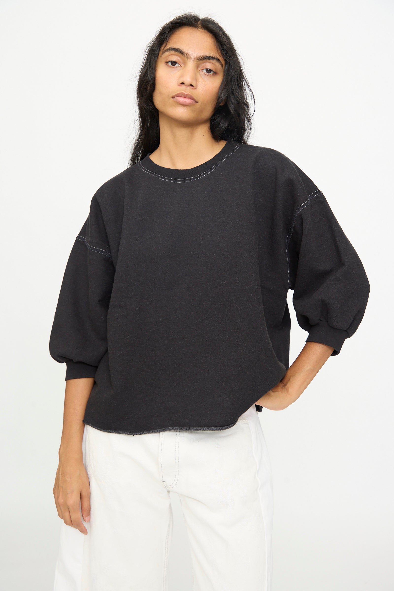 A person with long dark hair wears the Rachel Comey Fond Sweatshirt in Charcoal, an oversized style featuring balloon sleeves and a raw edge hemline, paired with white jeans and posed against a plain light background. The subtle detailing of the sweatshirt adds a modern touch to the relaxed look.