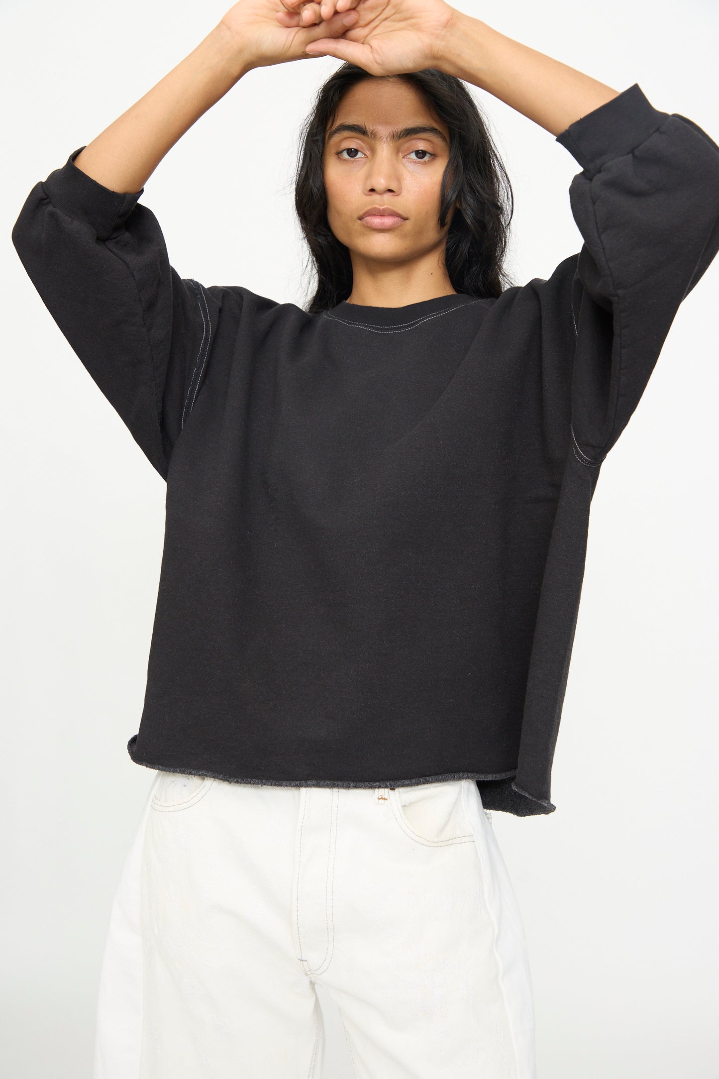 A person wearing the Rachel Comey Fond Sweatshirt in Charcoal, featuring an oversized fit and raw edge hemline, stands against a plain background with arms raised above their head, paired with white pants.