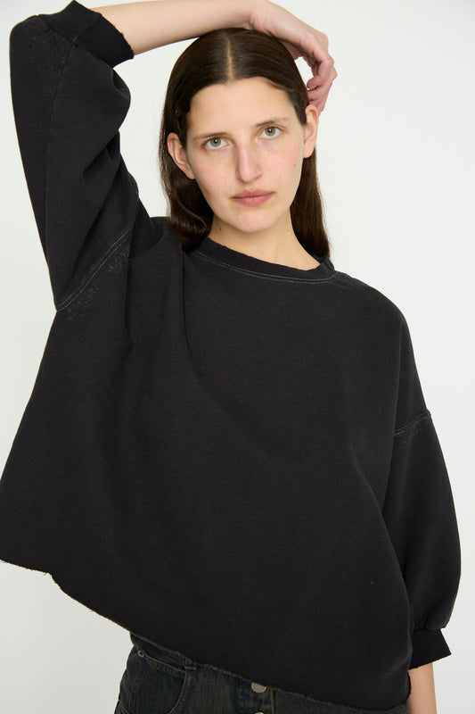 With long dark hair, a person in a Rachel Comey Fond Sweatshirt in Charcoal poses against a plain backdrop, raising an arm to touch their head. 