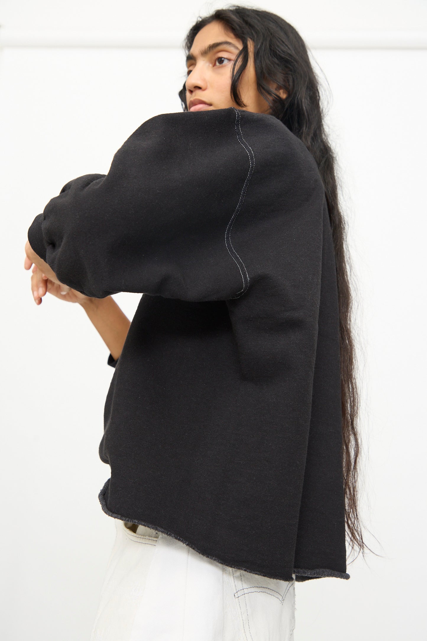 A person with long, dark hair wears the Rachel Comey Fond Sweatshirt in Charcoal—an oversized style featuring a raw edge hemline—paired with light pants. They stand against a plain white background, facing slightly away from the camera.