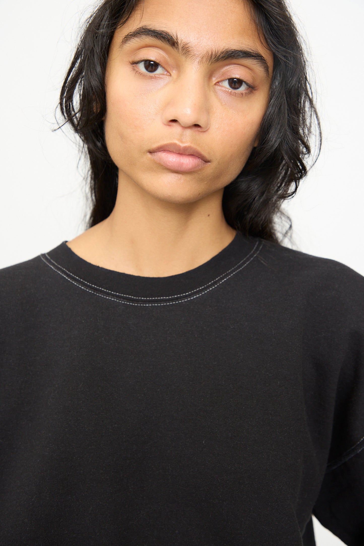 A person with long dark hair, wearing the Rachel Comey Fond Sweatshirt in Charcoal—an oversized style featuring a raw edge hemline—stands against a plain light background, gazing calmly at the camera.