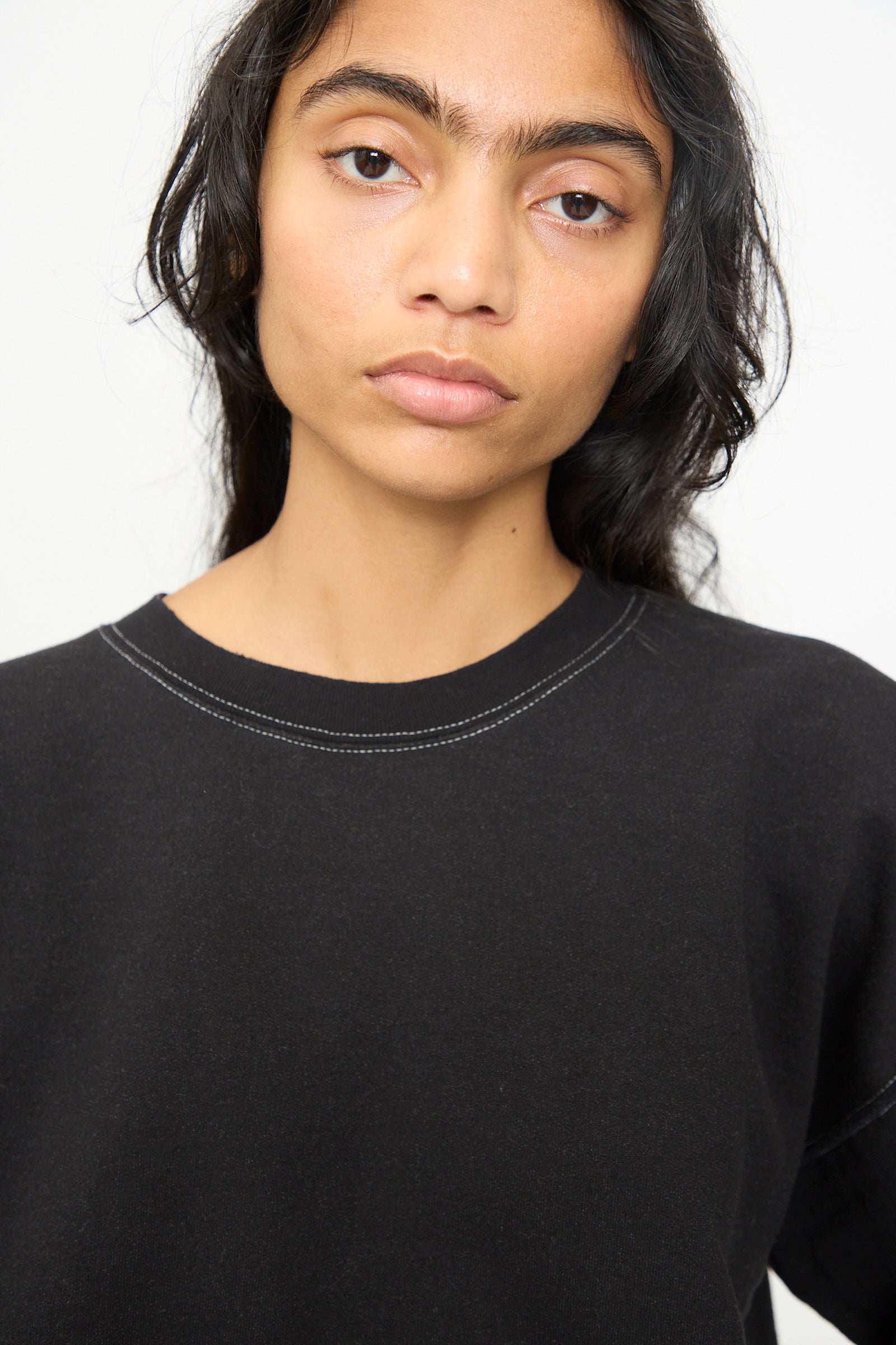 A person with long dark hair, wearing the Rachel Comey Fond Sweatshirt in Charcoal—an oversized style featuring a raw edge hemline—stands against a plain light background, gazing calmly at the camera.