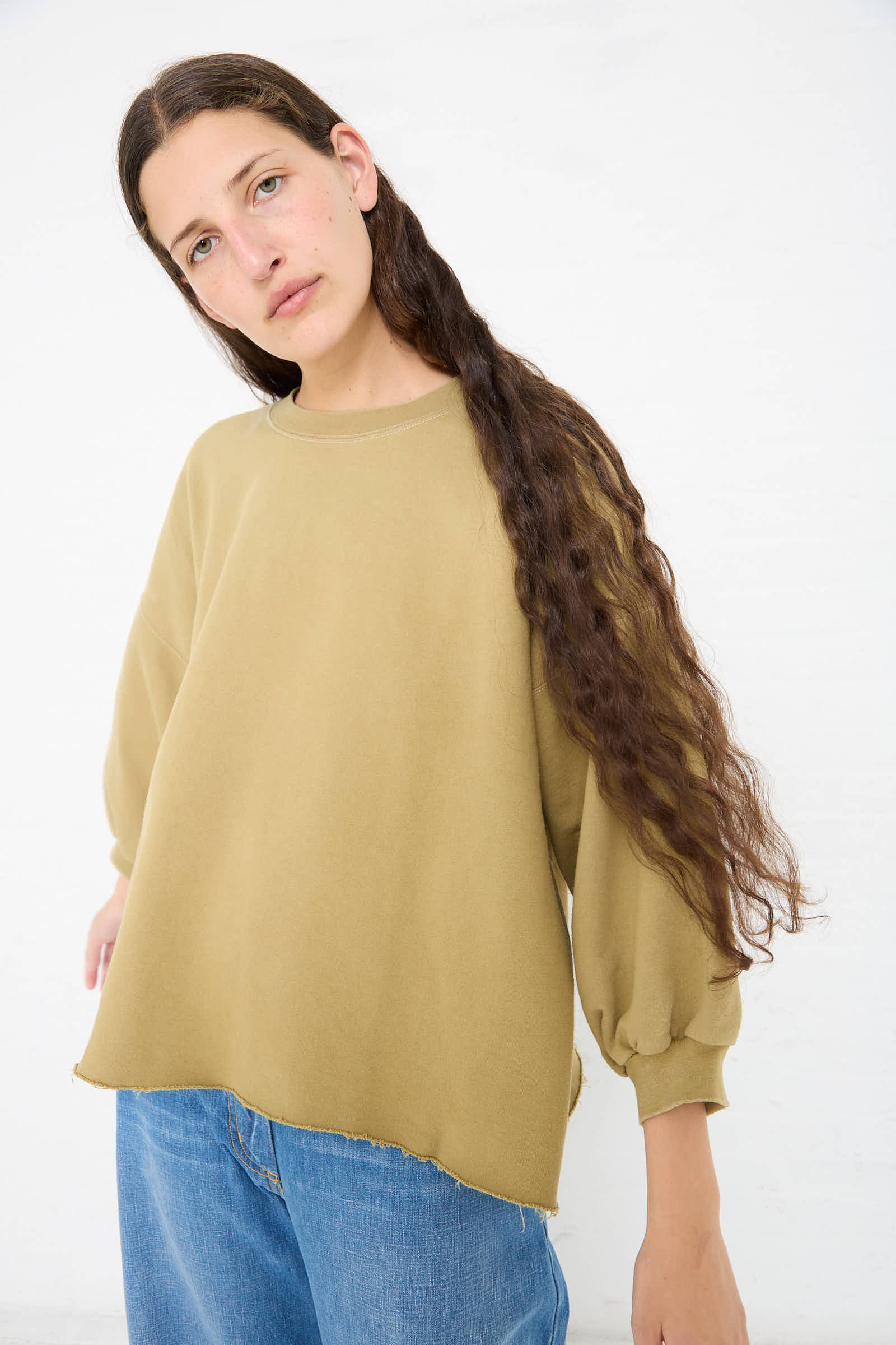 A person with long brown hair wearing a Rachel Comey Fond Sweatshirt in Dusty Sage and blue jeans stands against a plain white background.