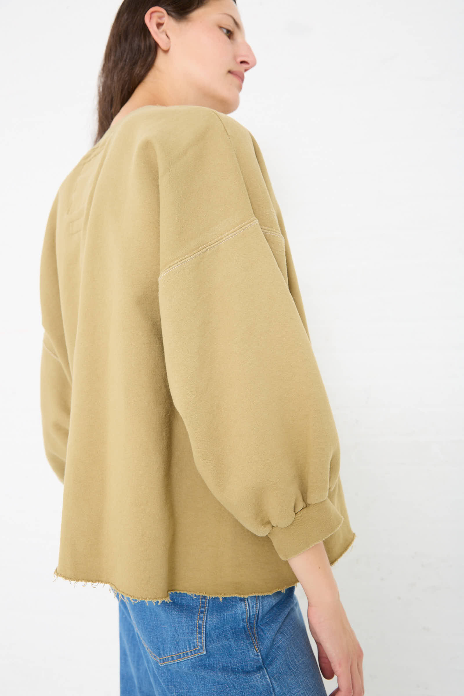 Person wearing a loose-fitting beige Fond Sweatshirt in Dusty Sage by Rachel Comey with voluminous cropped sleeves and blue jeans, standing against a plain white background.