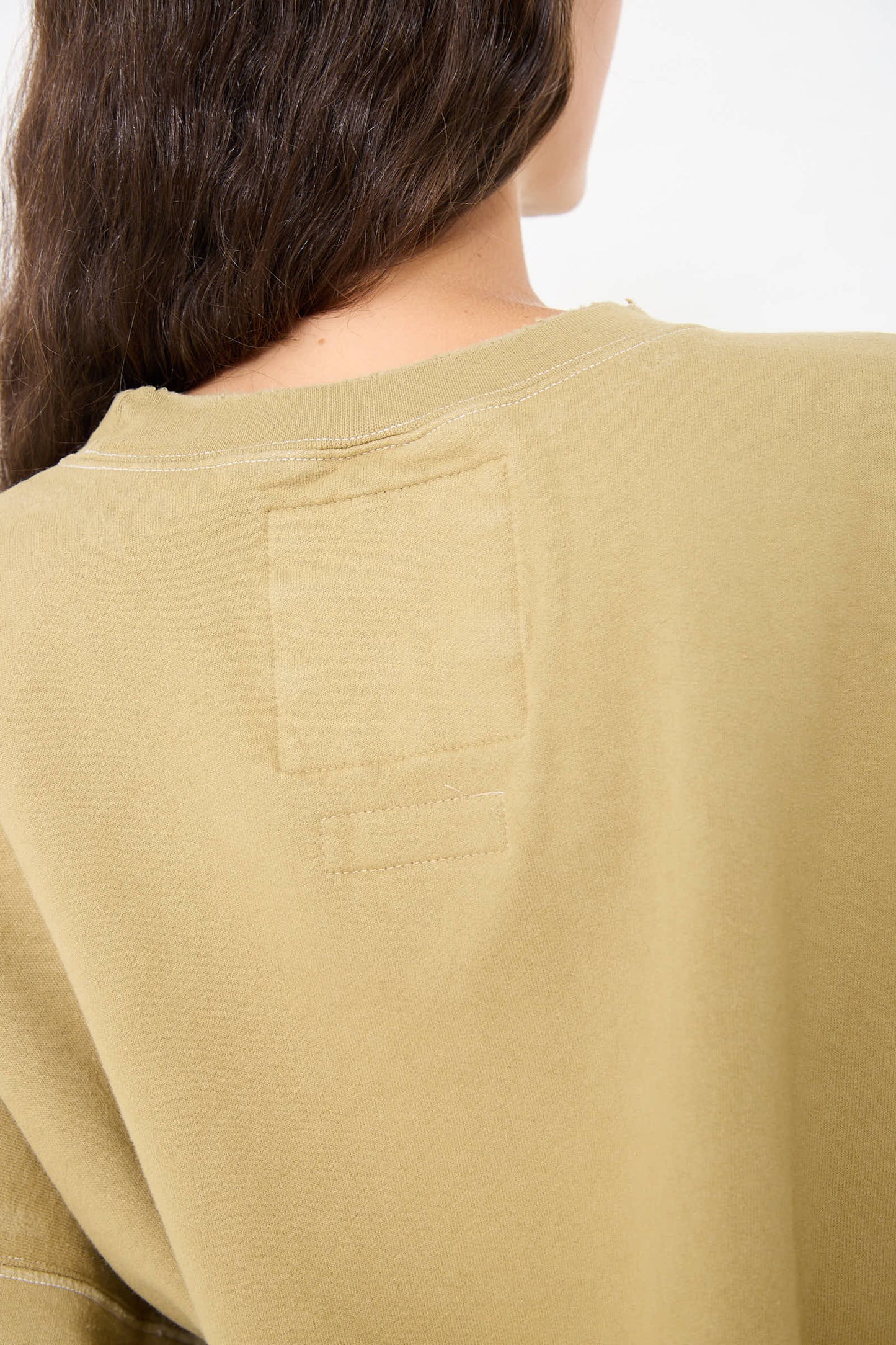 Close-up of the back of a person's head and shoulders, showing long brown hair cascading over a tan-colored Fond Sweatshirt in Dusty Sage by Rachel Comey with visible stitching and a rectangular patch near the collar.