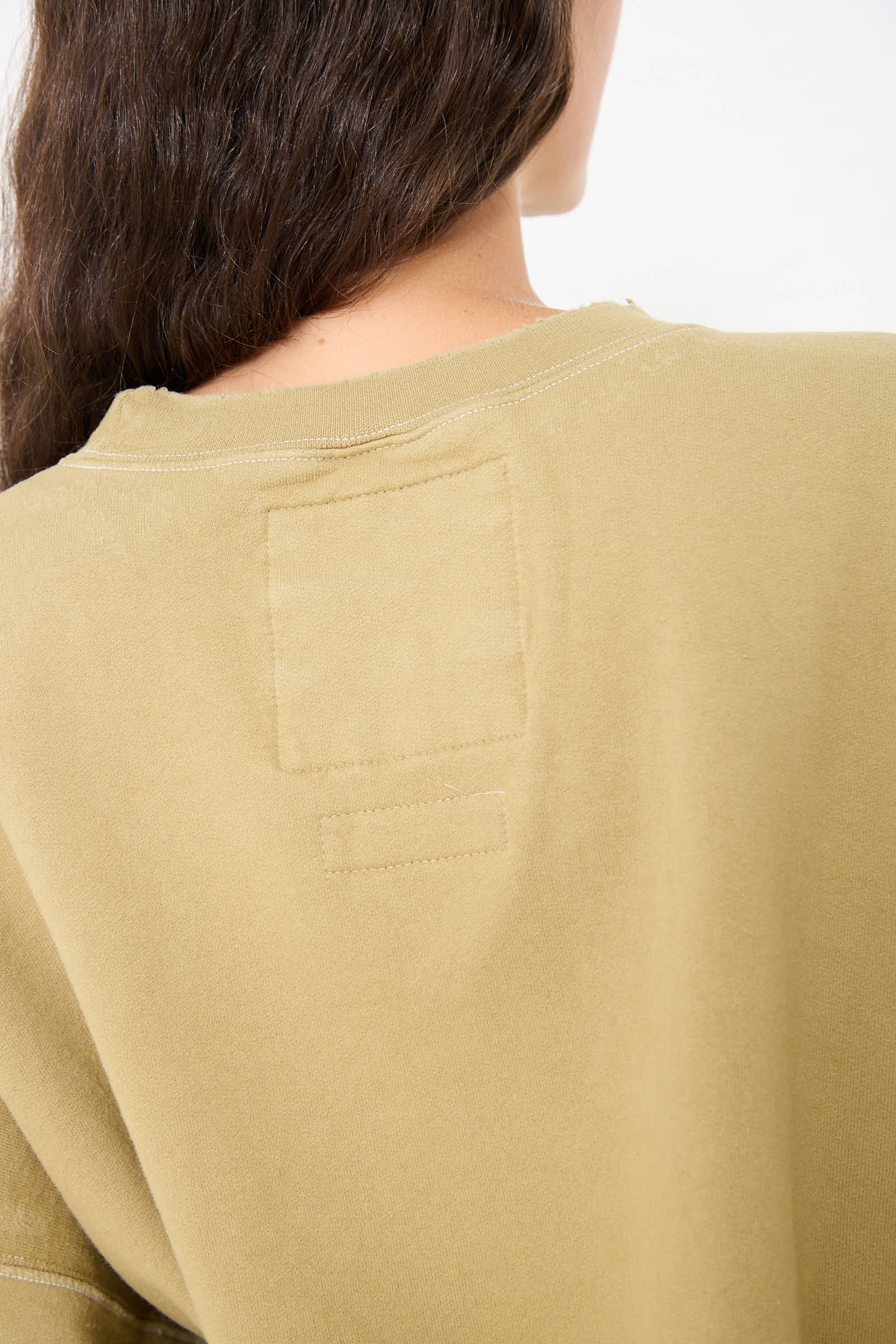 Close-up of the back of a person's head and shoulders, showing long brown hair cascading over a tan-colored Fond Sweatshirt in Dusty Sage by Rachel Comey with visible stitching and a rectangular patch near the collar.