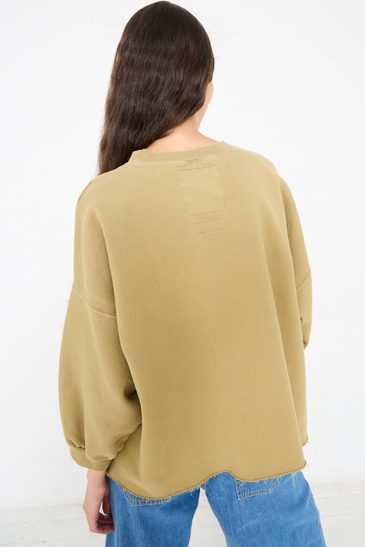 A person with long hair is seen from the back wearing a loose-fitting, oversized Rachel Comey Fond Sweatshirt in Dusty Sage with voluminous cropped sleeves and blue jeans.