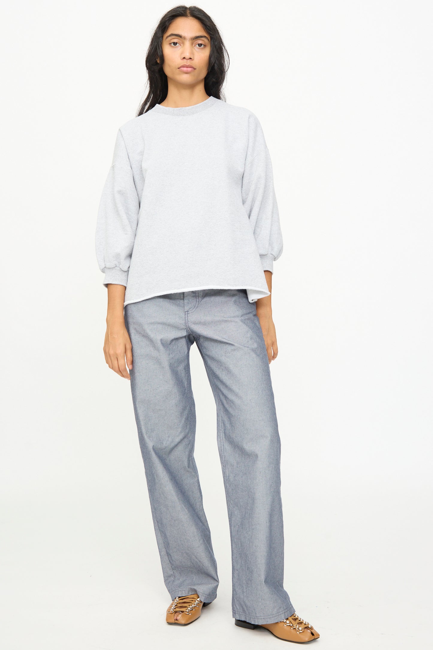 A person stands facing forward against a plain white background, wearing the Rachel Comey Fond Sweatshirt in Grey—an oversized piece featuring balloon sleeves and a raw edge hemline—paired with wide-leg gray pants and brown loafers.