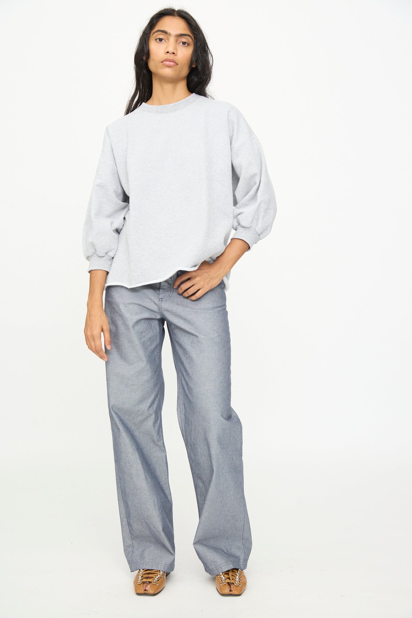 A person stands against a plain white background wearing the Rachel Comey Fond Sweatshirt in Grey, which features an oversized fit and raw edge hemline, paired with wide-leg blue pants and brown shoes, with one hand in their pocket.