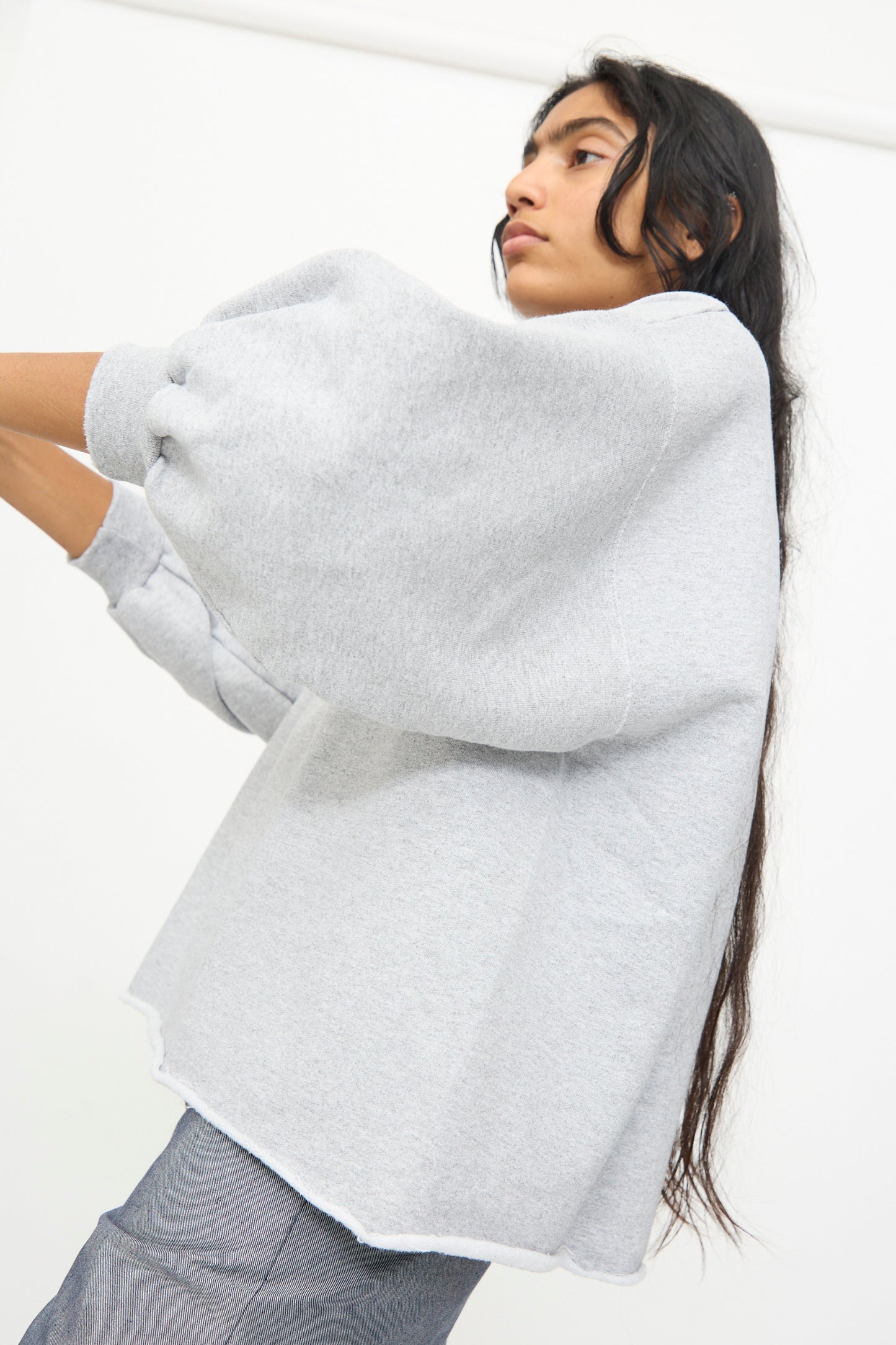 A person with long dark hair wears the Rachel Comey Fond Sweatshirt in Grey, featuring an oversized fit, voluminous sleeves, and a raw edge hemline, paired with gray pants and standing against a plain white background.