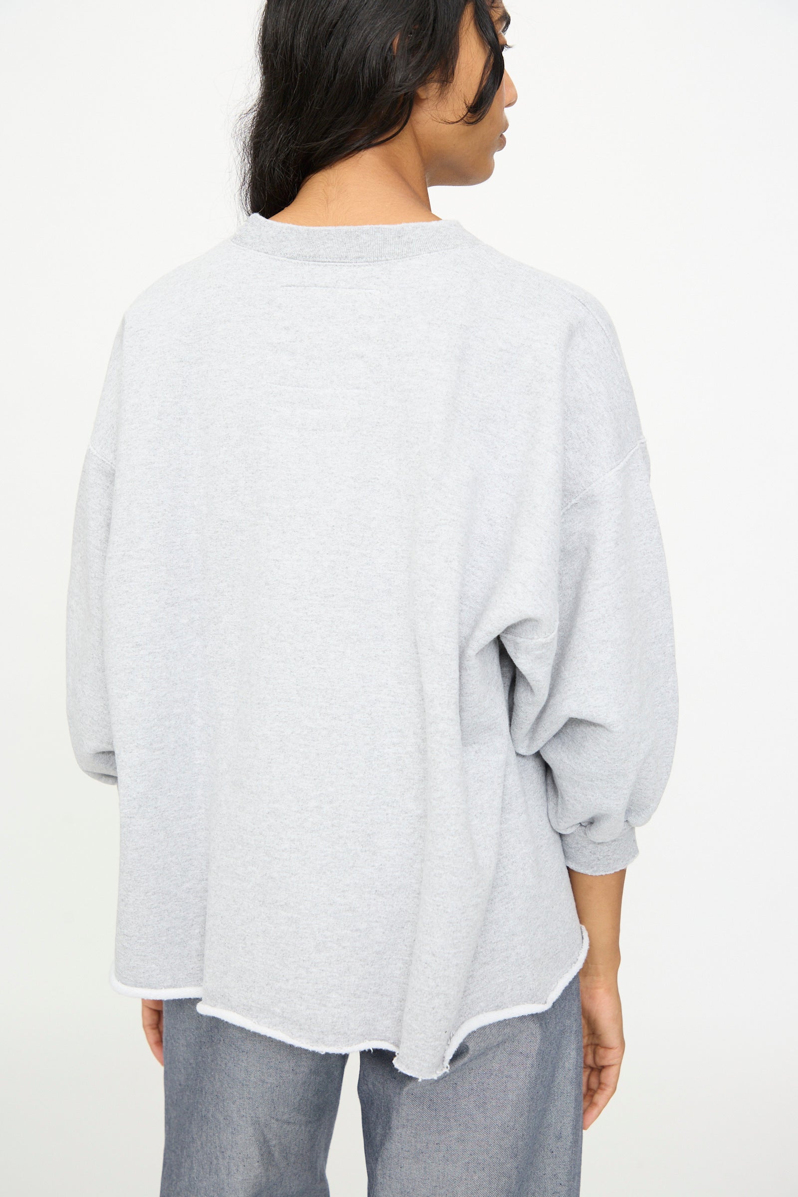 A person stands facing away, wearing the Rachel Comey Fond Sweatshirt in Grey—an oversized, loose-fitting piece with dropped shoulders, long sleeves, and a raw edge hemline—paired with gray pants.