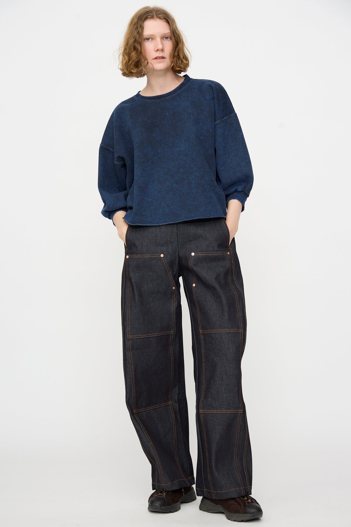 A person stands against a plain white background, wearing the Rachel Comey Fond Sweatshirt in Indigo—an oversized piece crafted from distressed cotton—paired with wide-leg indigo denim jeans featuring visible seams, hands in pockets, and black shoes.