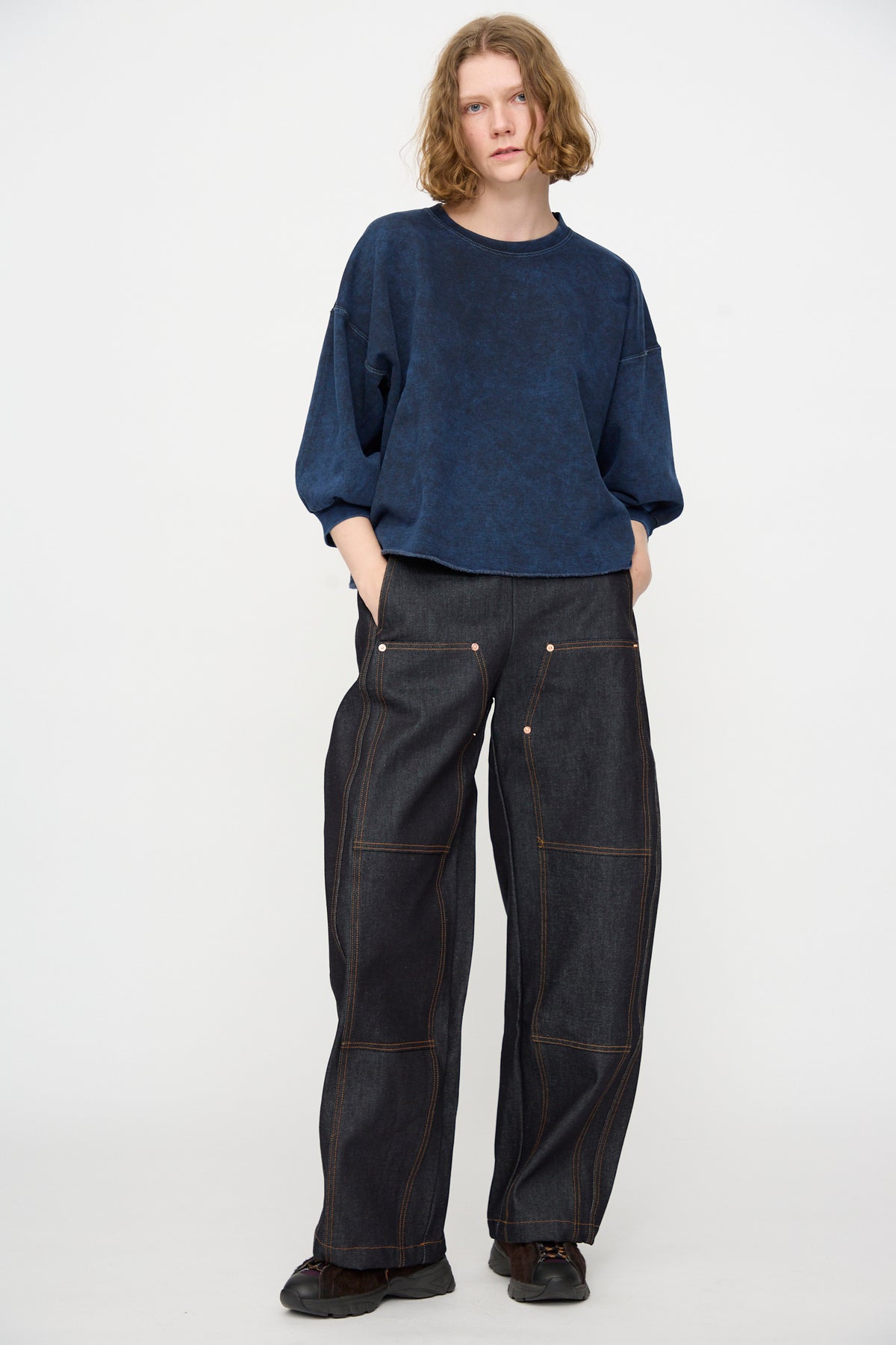 A person stands against a plain background, wearing the Rachel Comey Fond Sweatshirt in Indigo—an oversized, loose dark blue cotton top—paired with wide-leg dark denim jeans featuring visible stitching and black shoes. Their hands are in their pockets and their expression is neutral.