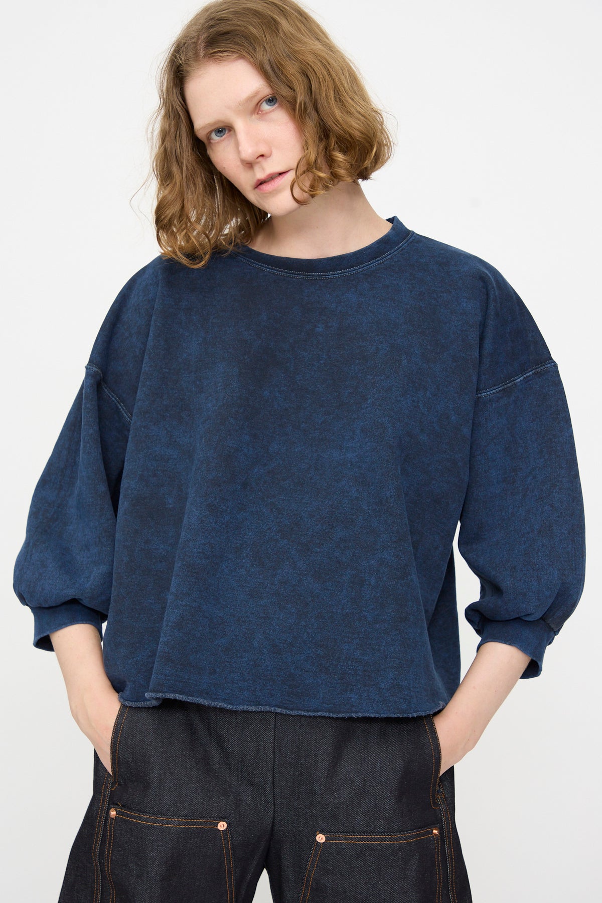 A person with shoulder-length curly hair wears the Rachel Comey Fond Sweatshirt in Indigo—an oversized, distressed cotton top—paired with dark pants featuring front pockets, standing against a plain light background.