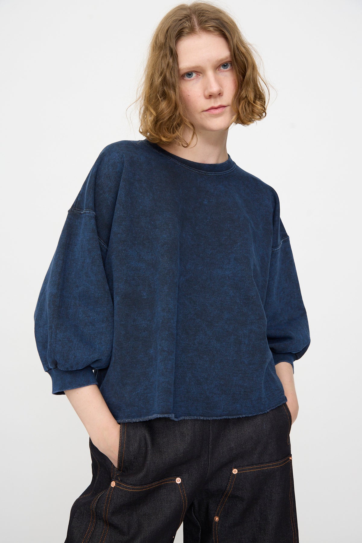 A person with shoulder-length wavy hair wears the Rachel Comey Fond Sweatshirt in Indigo—an oversized, distressed cotton piece—paired with dark pants, standing against a plain white background with hands in pockets.