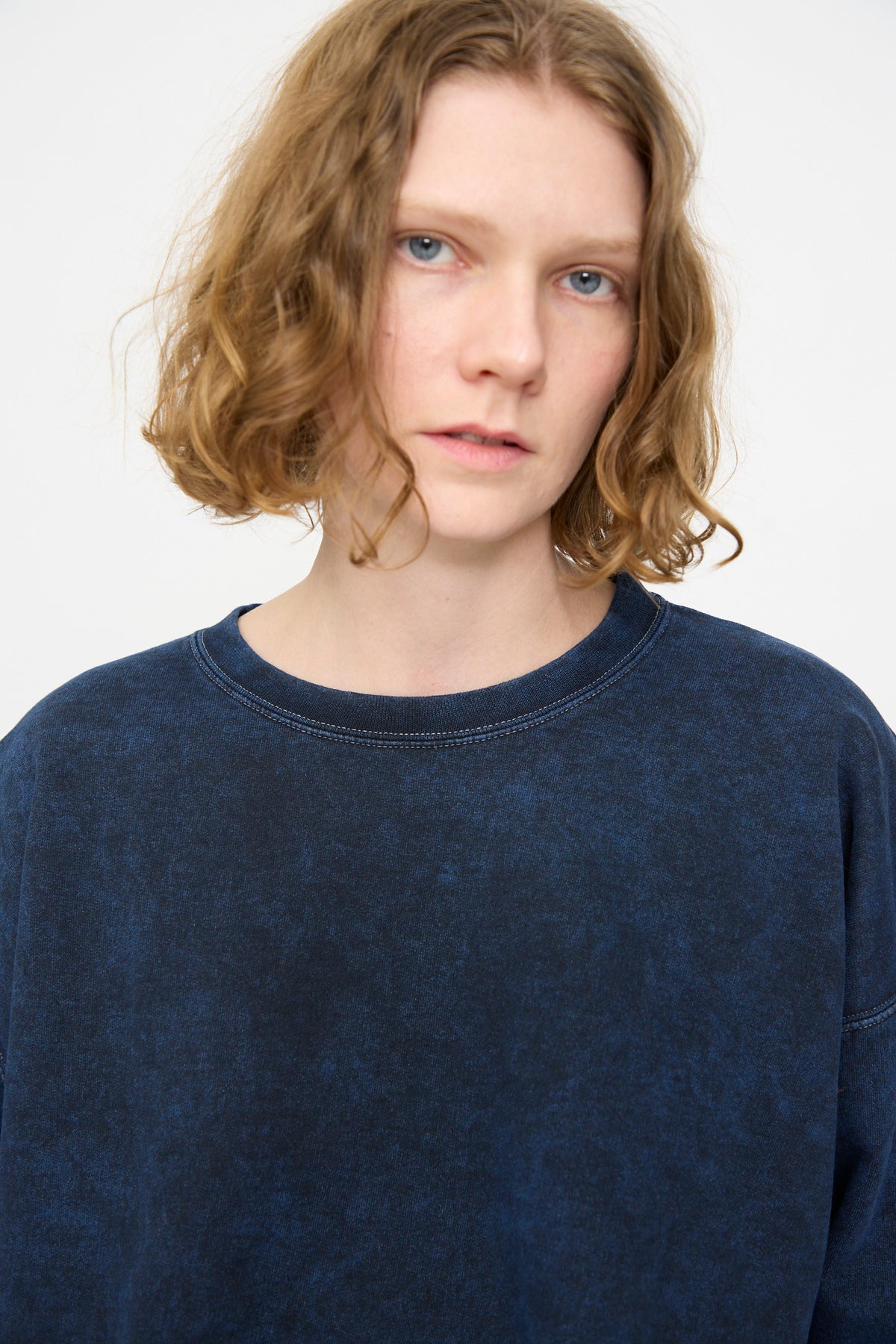 A person with short, wavy, light brown hair wears the Rachel Comey Fond Sweatshirt in Indigo—an oversized crewneck crafted from distressed cotton—while looking directly at the camera with a neutral expression against a plain background.