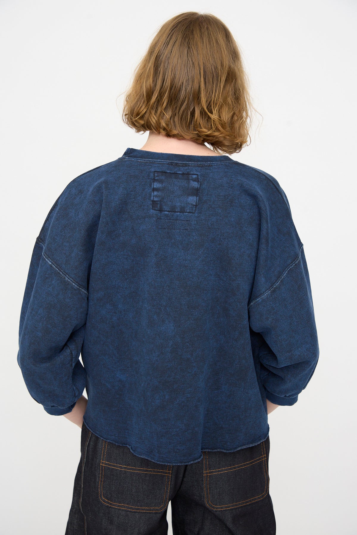 A person with shoulder-length light brown hair faces away, wearing the Rachel Comey Fond Sweatshirt in Indigo—an oversized style crafted from distressed cotton—paired with dark pants against a plain white background.