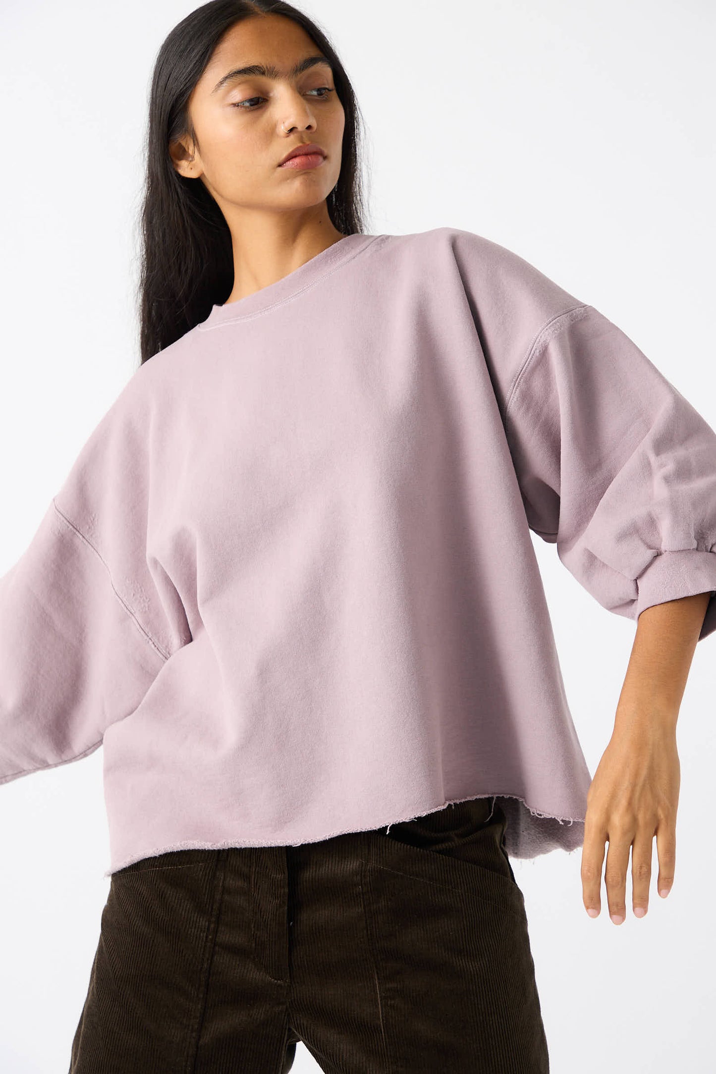 A person with long hair stands against a plain white background, wearing the Rachel Comey Fond Sweatshirt in Mauve and dark-colored pants. The sweatshirt features an oversized fit and a raw edge hemline, adding a touch of casual flair.