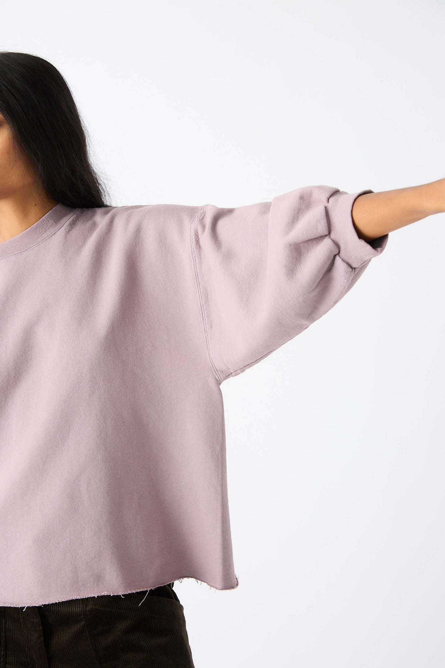 A person with long dark hair, wearing the Rachel Comey Fond Sweatshirt in Mauve, extends their right arm sideways against a plain background. The image is cropped at the shoulders.