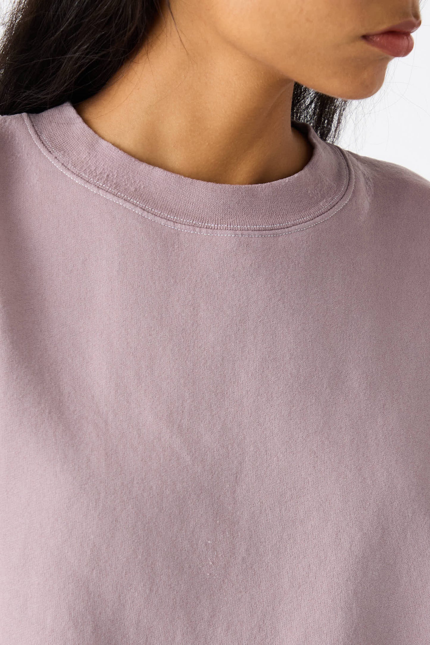 Close-up of a person wearing the Fond Sweatshirt in Mauve by Rachel Comey. The genderless, oversized sweatshirt features a raw edge hemline. The individual’s dark hair is partly visible, with the photo cropped from the nose down to the upper chest, showcasing the casual and relaxed vibe of this distressed cotton blend garment.