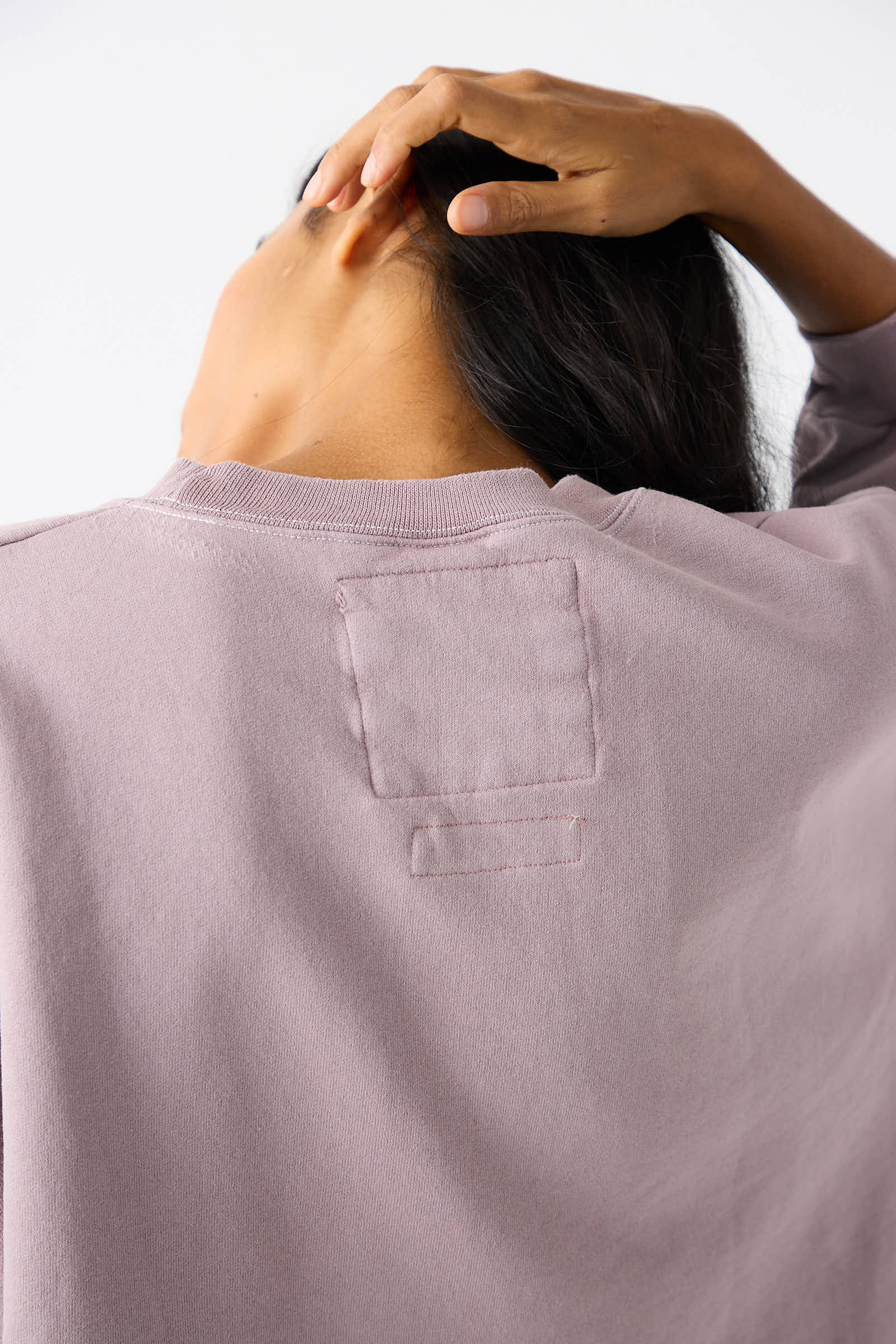 A person with long dark hair is shown from the back, wearing the Rachel Comey Fond Sweatshirt in Mauve, which features a stitched square patch on the upper back. The person's left hand is touching their head.