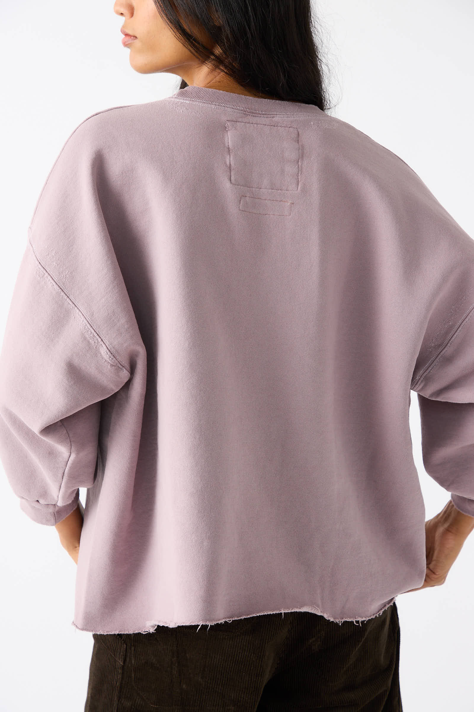 Person wearing an oversized, long-sleeved Rachel Comey Fond Sweatshirt in mauve with a raw edge hemline and dark trousers is shown from the back.