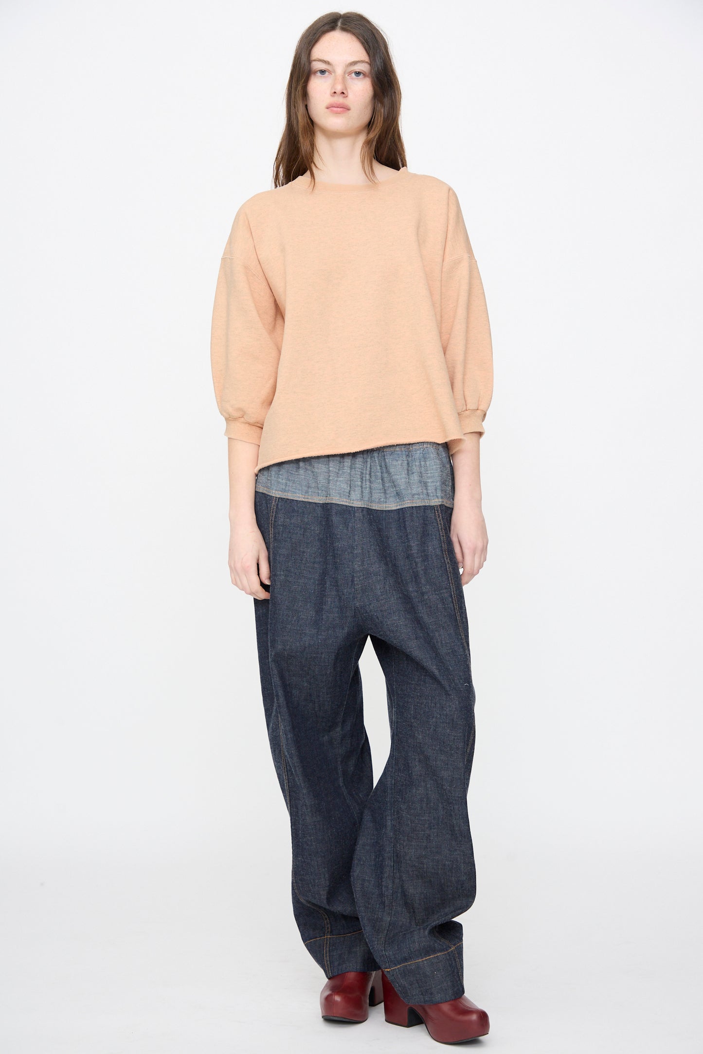 A person stands against a plain background wearing the Rachel Comey Fond Sweatshirt in Peach, styled with a gray undershirt, loose dark denim pants, and red shoes.
