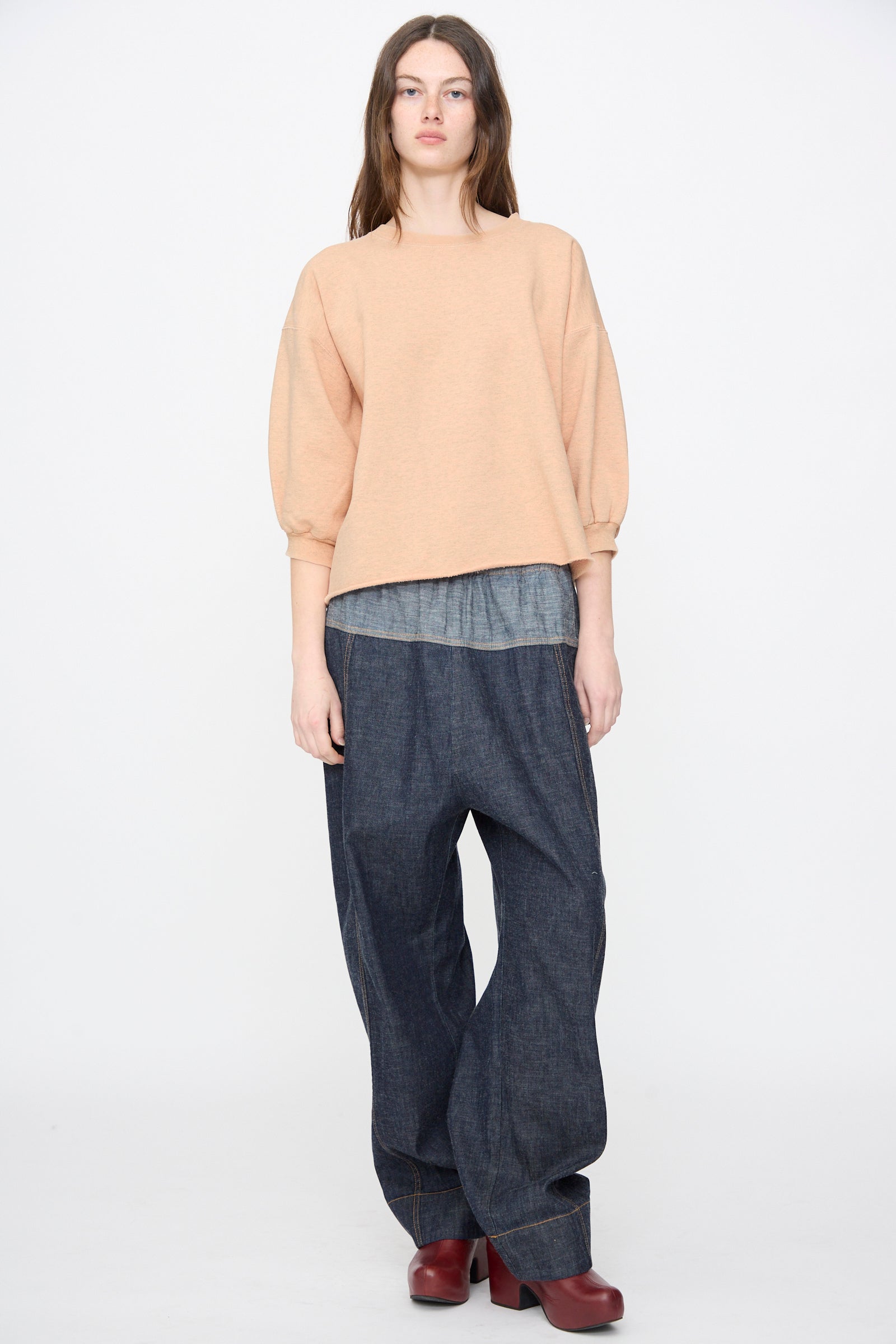A person stands against a plain background wearing the Rachel Comey Fond Sweatshirt in Peach, styled with a gray undershirt, loose dark denim pants, and red shoes.