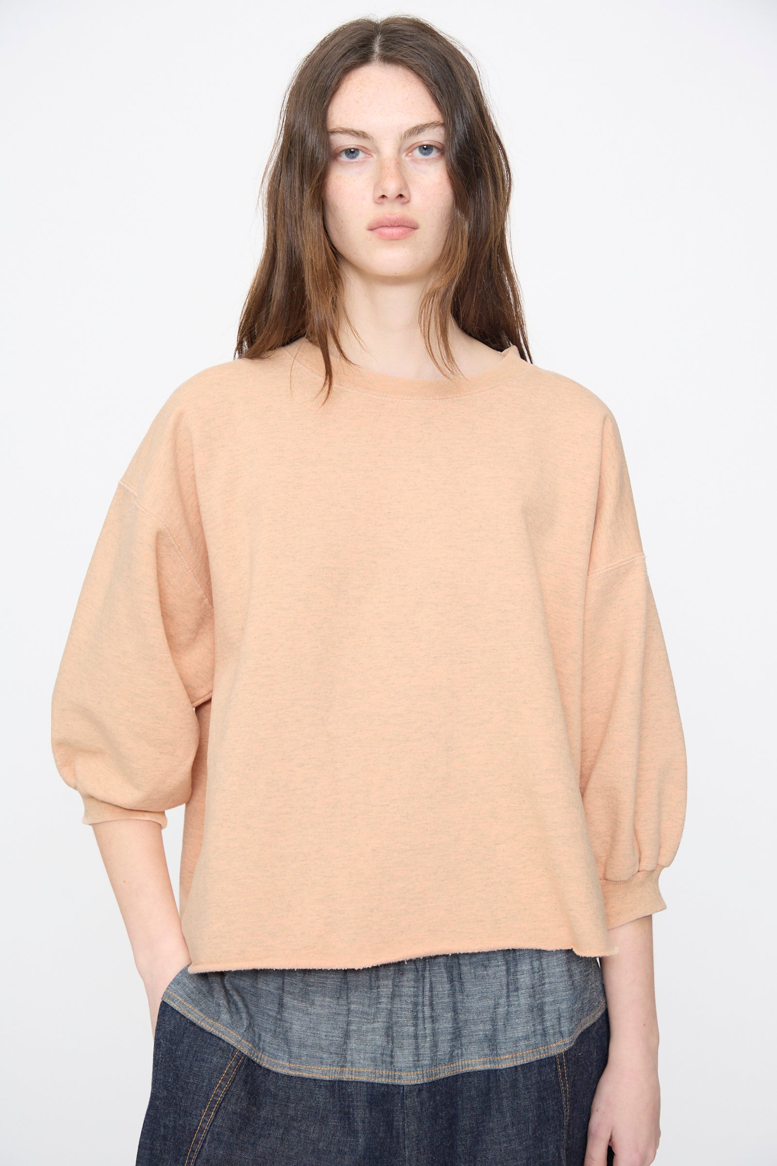 A person with long brown hair wears the Rachel Comey Fond Sweatshirt in Peach—an oversized, genderless top with three-quarter sleeves—paired with a dark denim skirt, standing against a plain background.