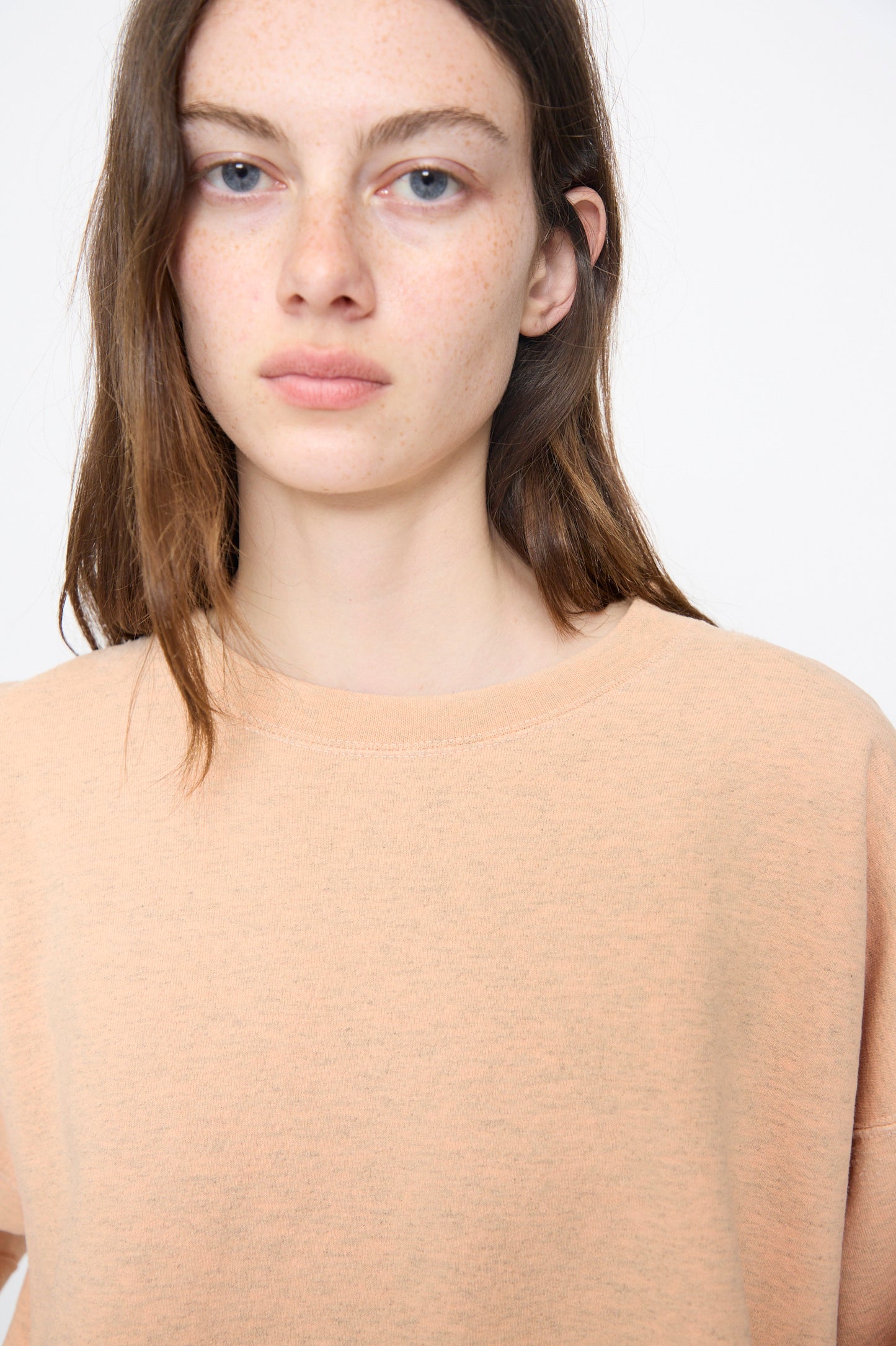 A young woman with long brown hair and freckles wears the Rachel Comey Fond Sweatshirt in Peach, a genderless oversized style with a relaxed fit and raw edge hemline, standing against a plain white background.