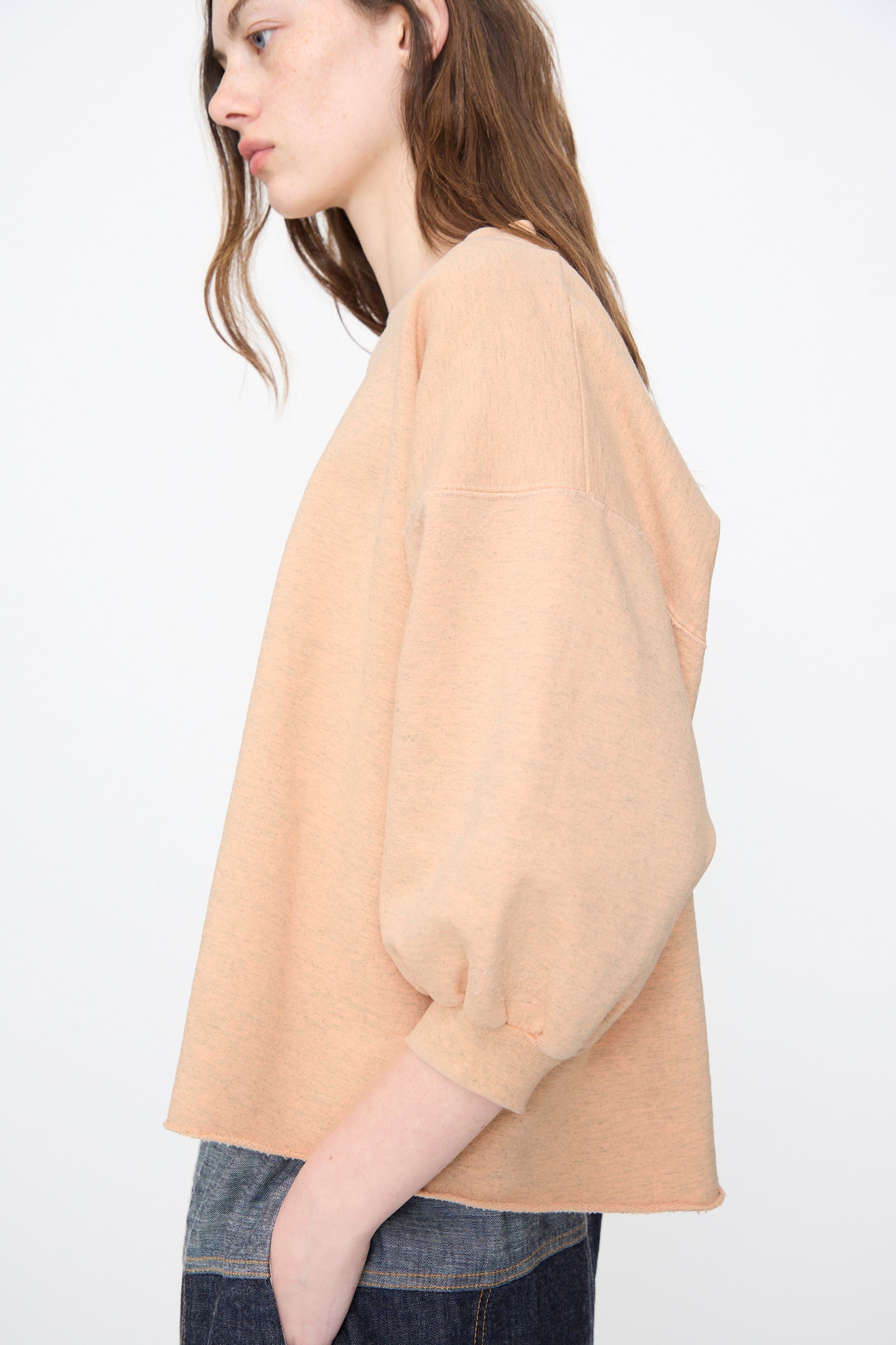 A person stands sideways against a plain light background wearing the Rachel Comey Fond Sweatshirt in Peach, a loose distressed cotton-blend long-sleeve top with balloon sleeves, paired with dark jeans.
