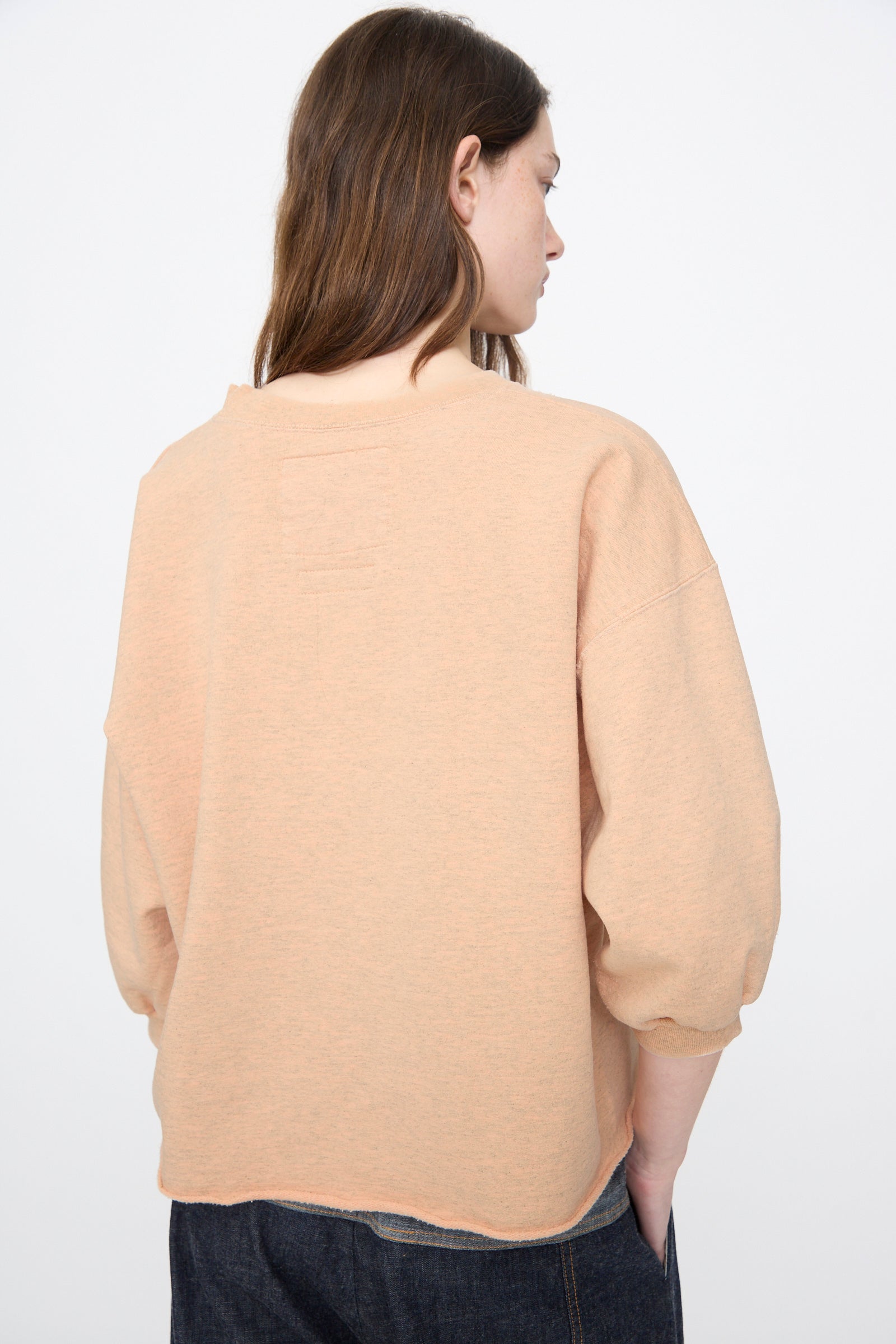 A woman with long brown hair stands with her back to the camera, wearing the Rachel Comey Fond Sweatshirt in Peach—a loose, distressed cotton blend with three-quarter sleeves—paired with dark jeans against a plain background.