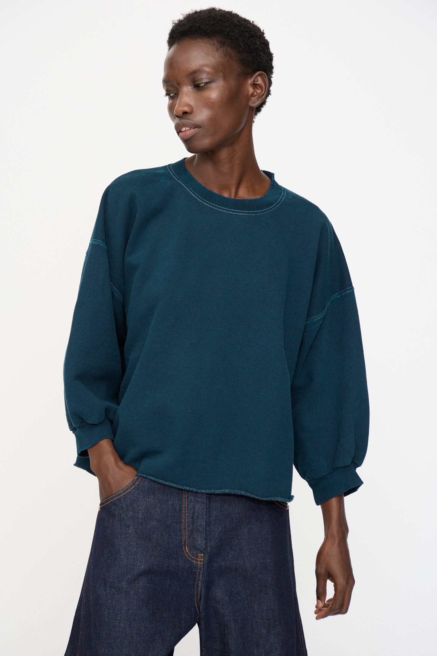 Model wears the Rachel Comey Fond Sweatshirt in Teal—an oversized, genderless piece with distressed cotton and three-quarter sleeves—paired with dark denim jeans, standing against a plain white background.