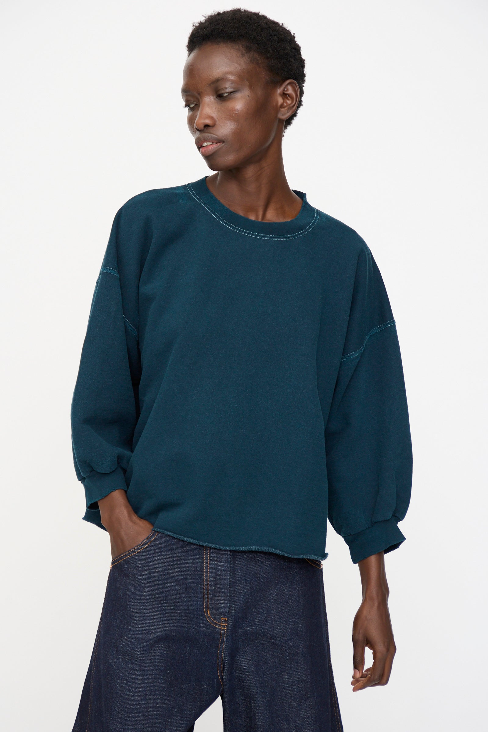 Model wears the Rachel Comey Fond Sweatshirt in Teal—an oversized, genderless piece with distressed cotton and three-quarter sleeves—paired with dark denim jeans, standing against a plain white background.