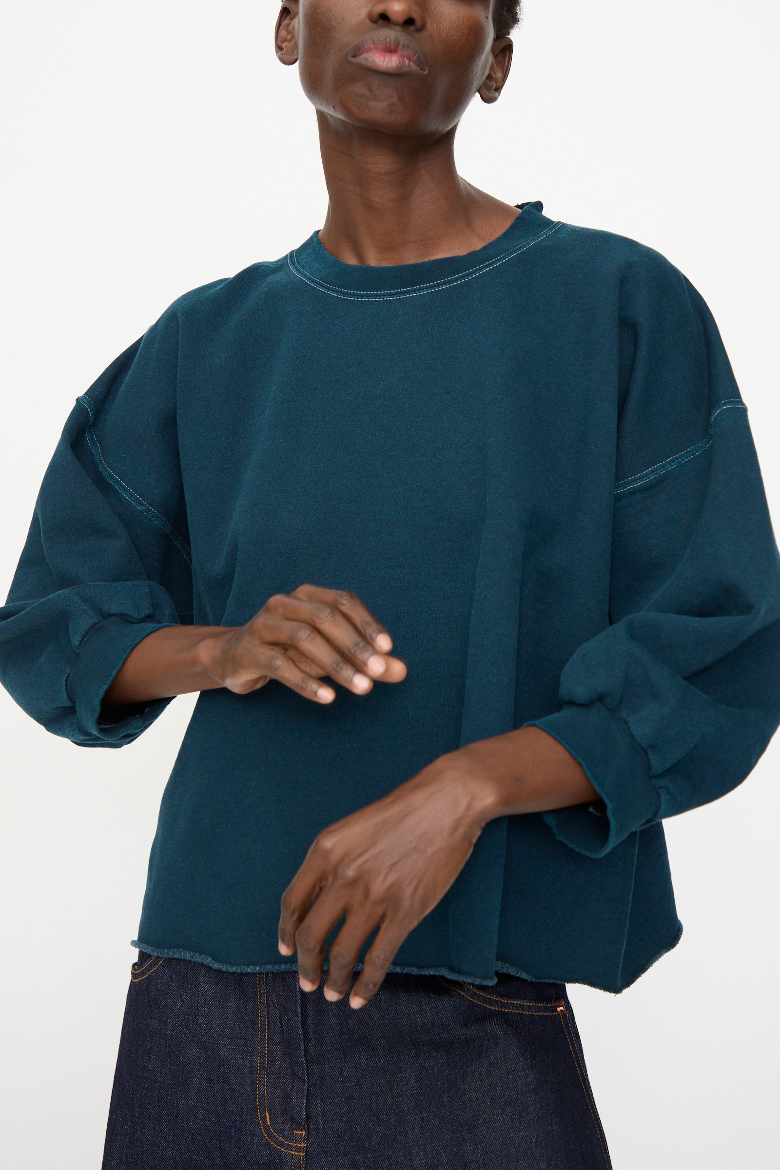 A person wearing the Rachel Comey Fond Sweatshirt in Teal, a loose genderless piece in distressed cotton, paired with dark denim pants, stands against a plain light background.