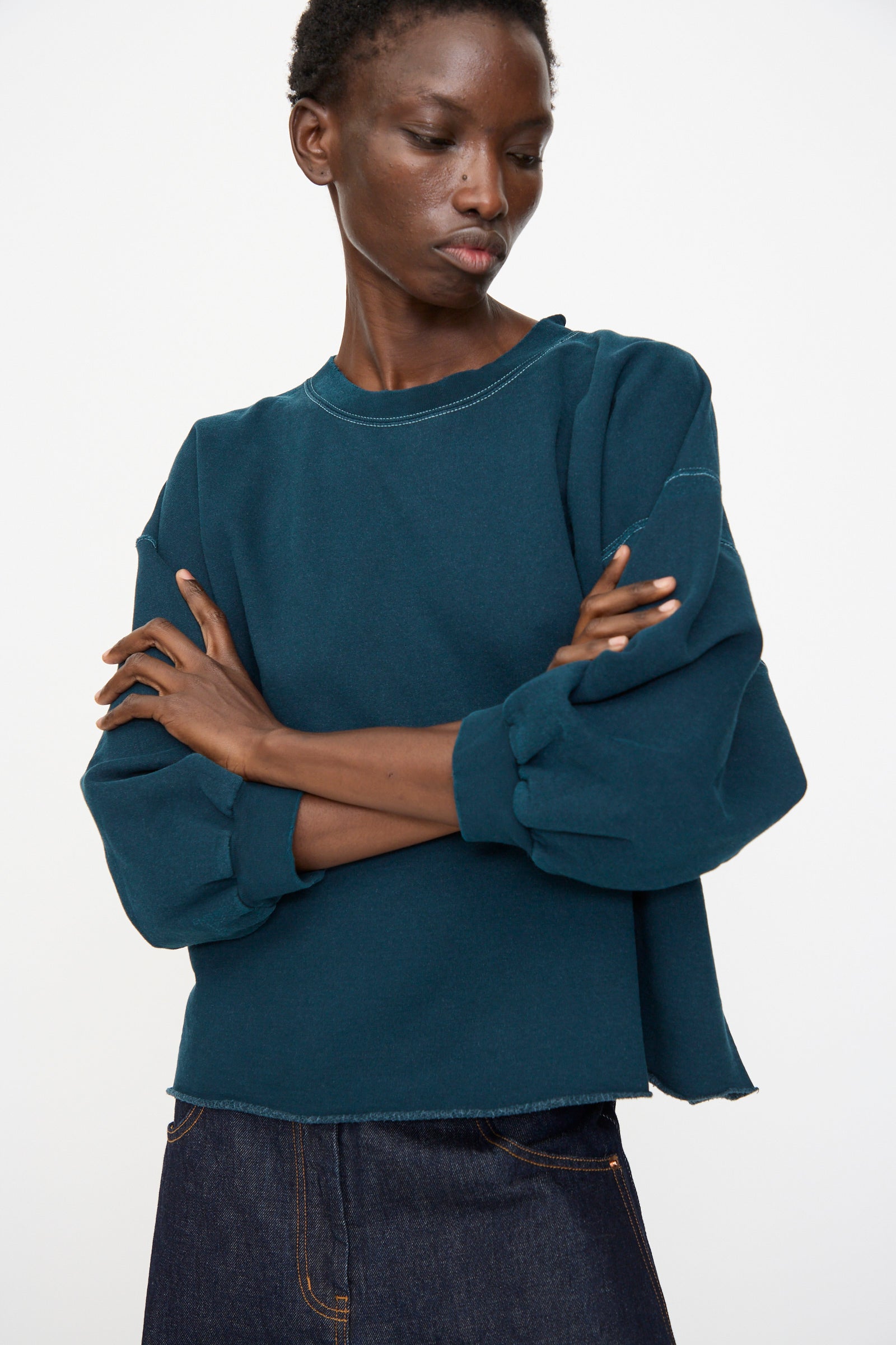 A person wearing the Rachel Comey Fond Sweatshirt in Teal with raw hems and dark jeans stands with arms crossed against a plain light background.