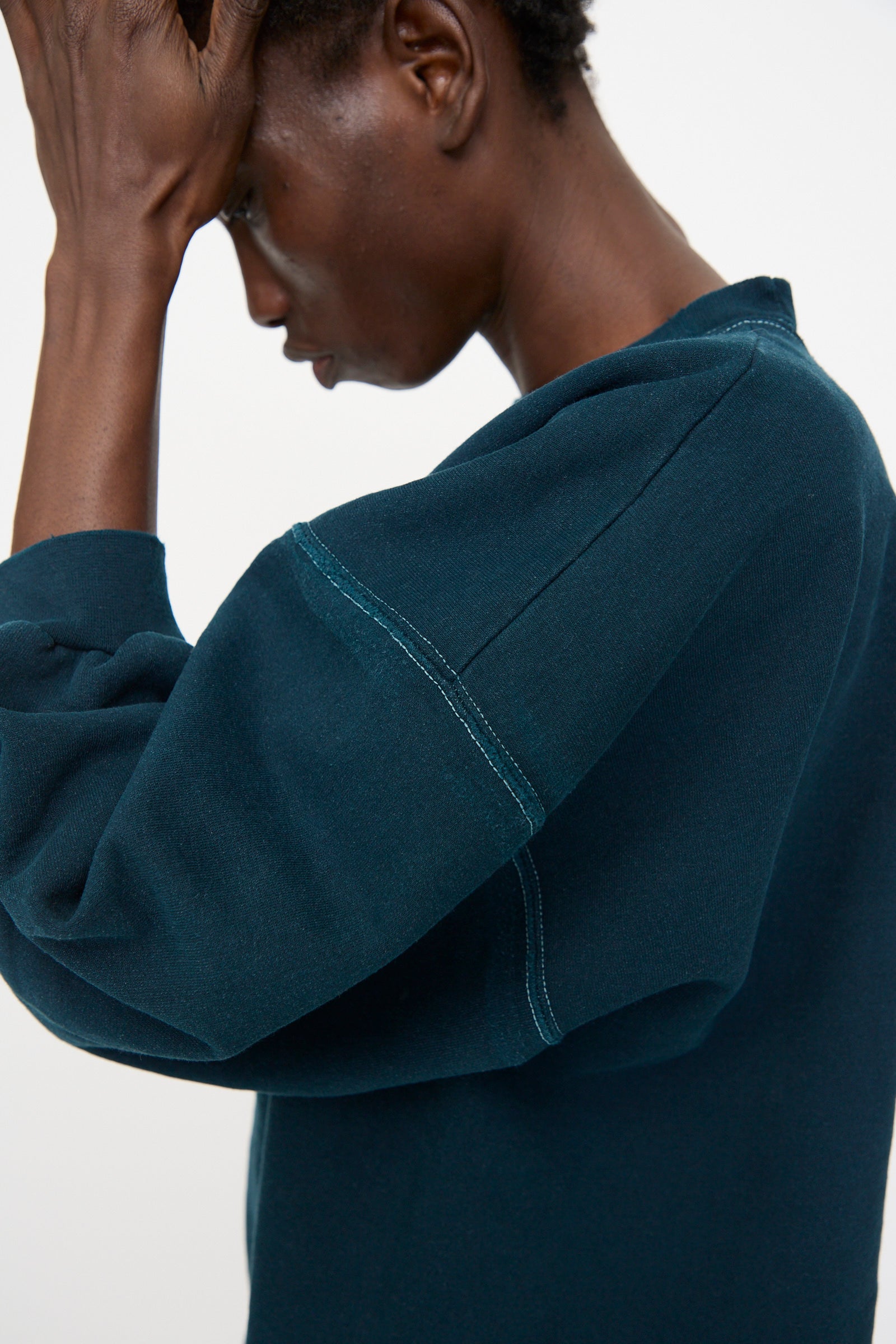 A person faces sideways, wearing the Rachel Comey Fond Sweatshirt in Teal—an oversized style with visible seam stitching—and holds their hand to their forehead.