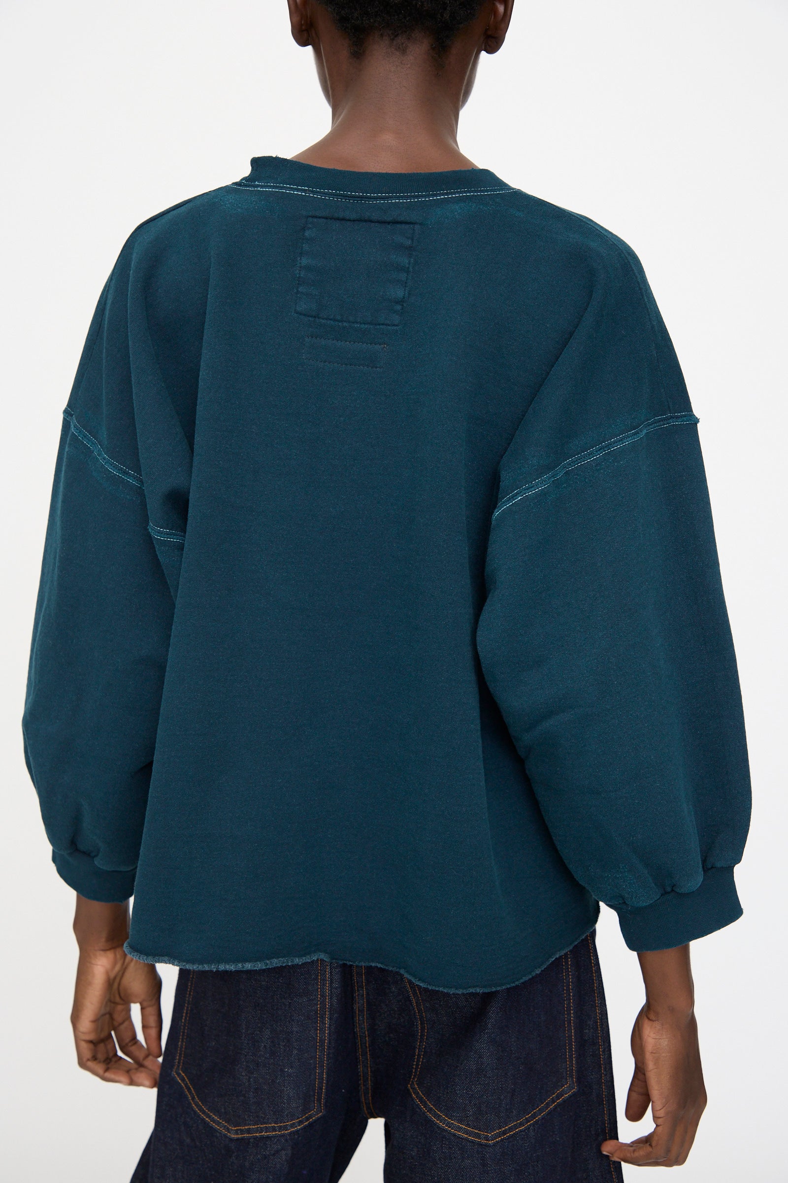 A person is seen from the back against a plain background, wearing the Rachel Comey Fond Sweatshirt in teal—an oversized style with balloon sleeves made from distressed cotton—paired with dark jeans.