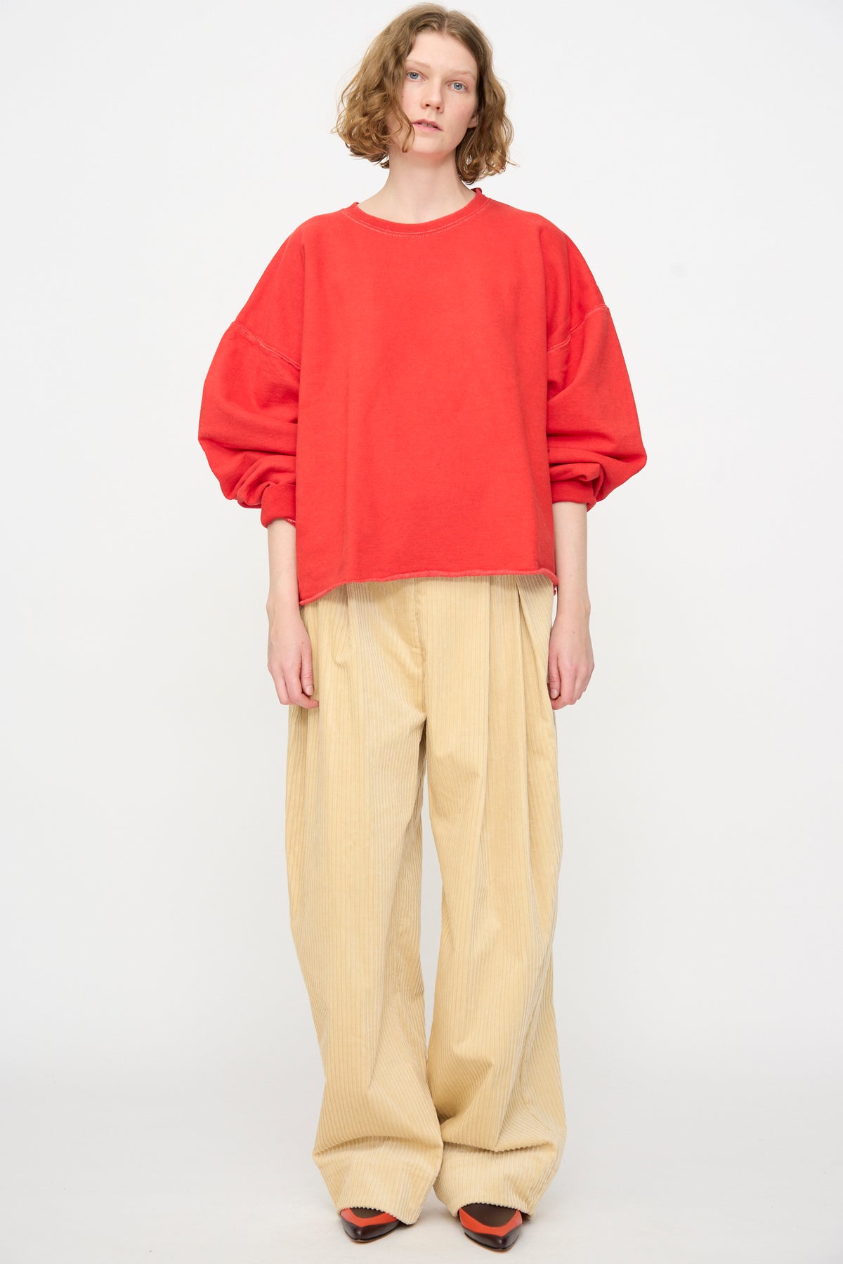 Standing against a plain background, the person is wearing the Rachel Comey Fonder Sweatshirt in Cherry, paired with wide beige pants and orange shoes.