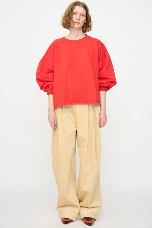 Standing against a plain background, the person is wearing the Rachel Comey Fonder Sweatshirt in Cherry, paired with wide beige pants and orange shoes.