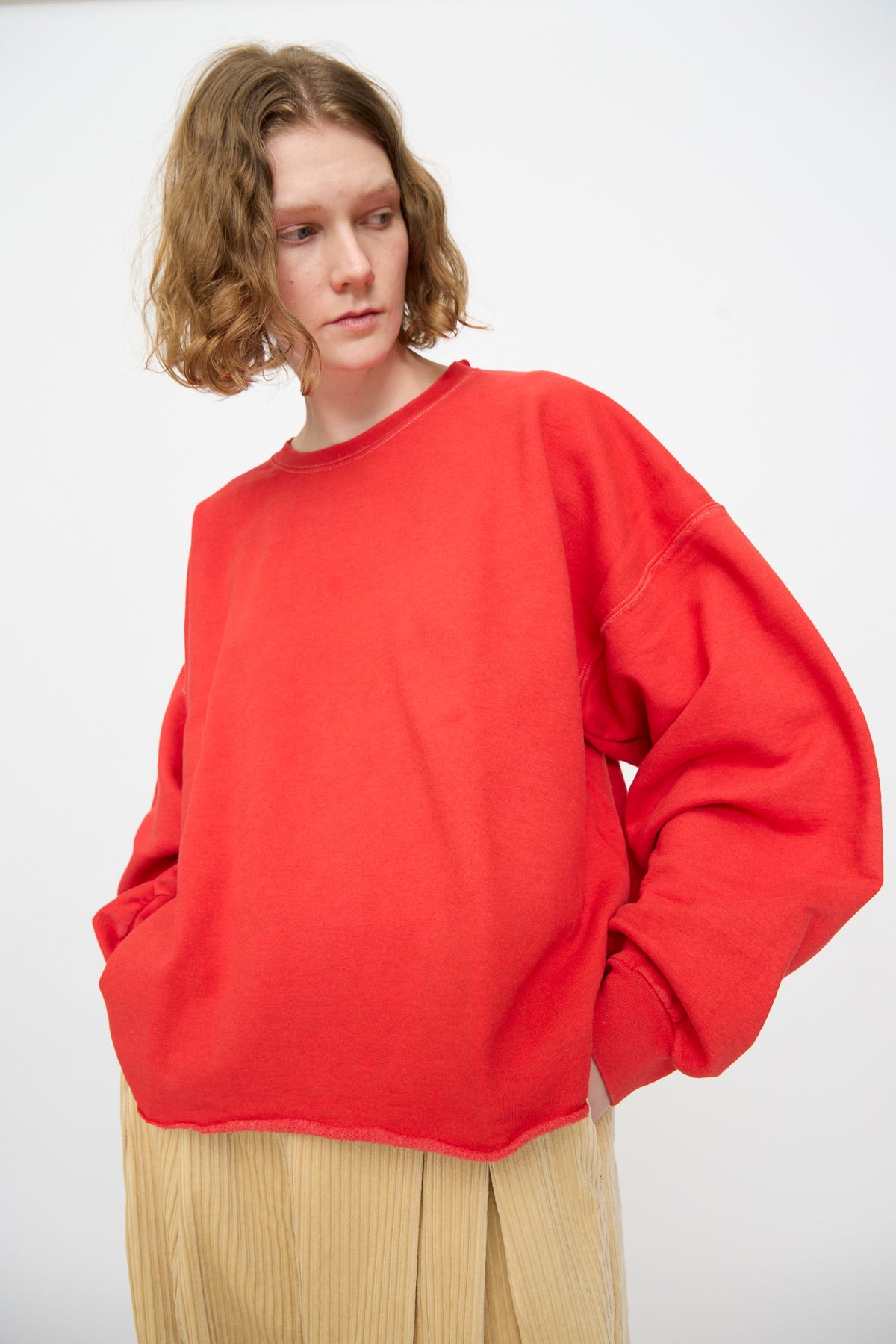 A person with wavy, shoulder-length hair wears the Rachel Comey Fonder Sweatshirt in Cherry and beige pleated pants, standing against a plain white background with hands in pockets, looking to the side.