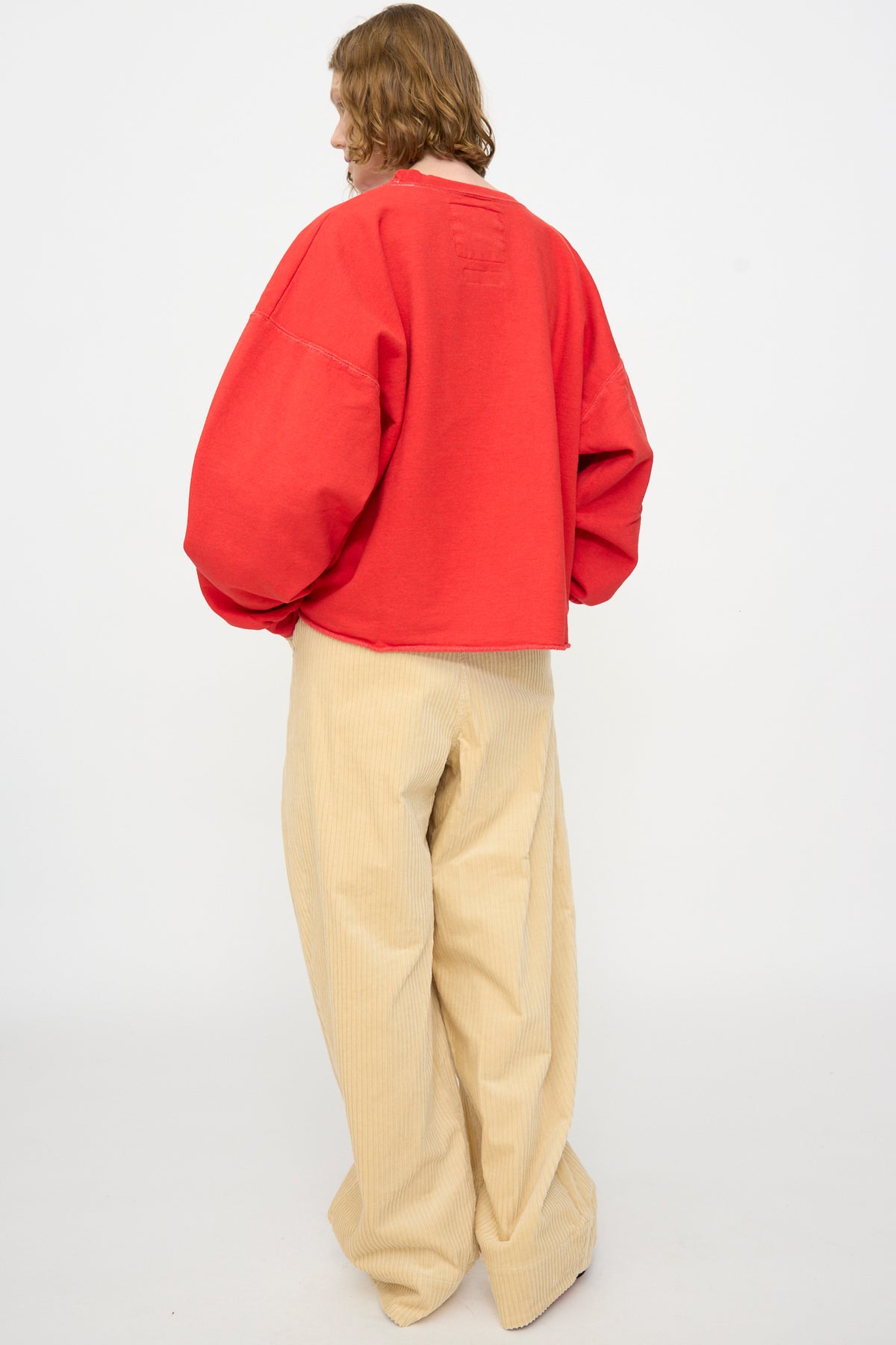 A person stands with their back to the camera, wearing the Rachel Comey Fonder Sweatshirt in Cherry—an oversized red sweatshirt with balloon sleeves—paired with wide-leg beige corduroy pants in a relaxed fit, set against a plain white background.