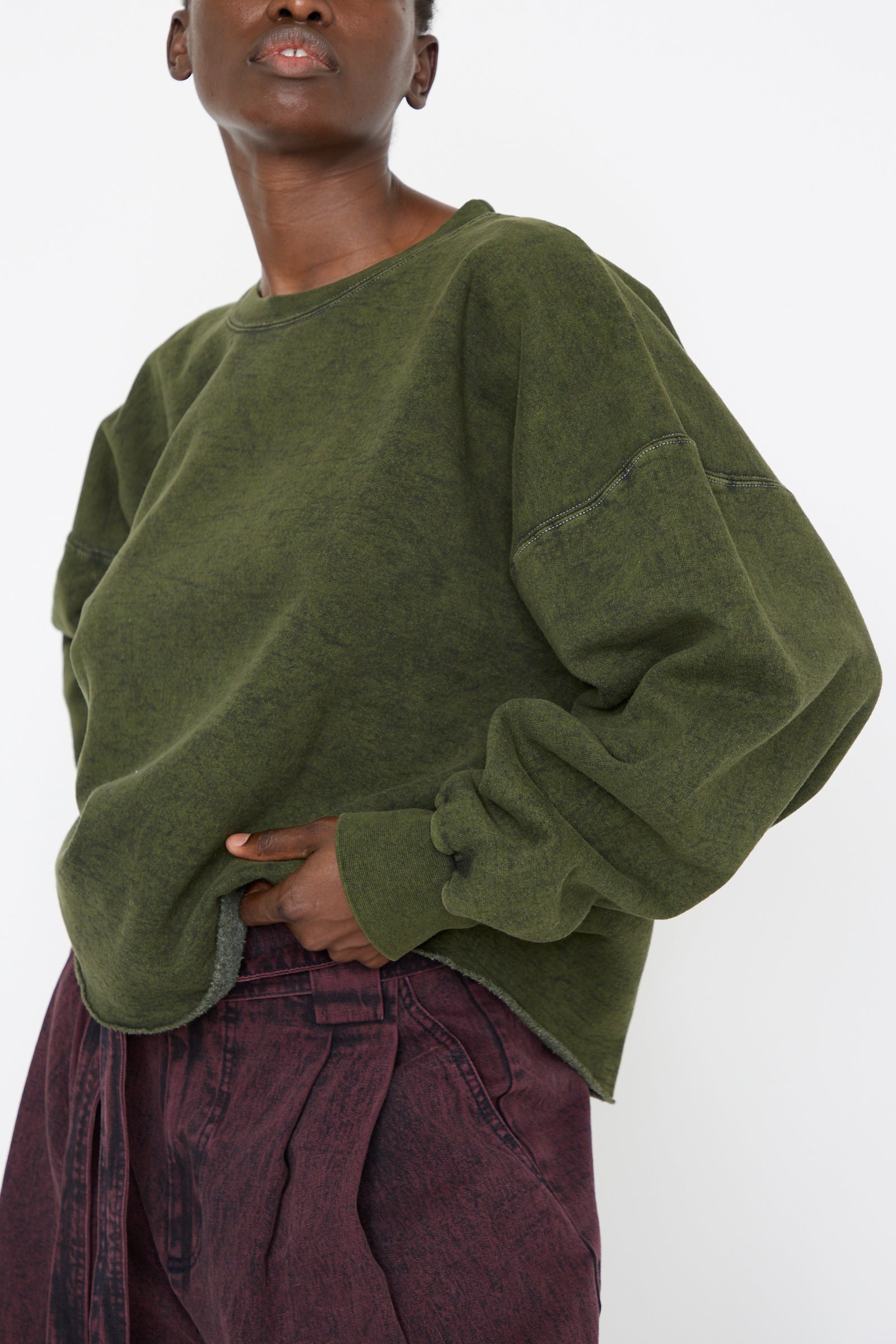 A woman wears the Fonder Sweatshirt in Olive by Rachel Comey, paired with faded maroon pants against a plain background.