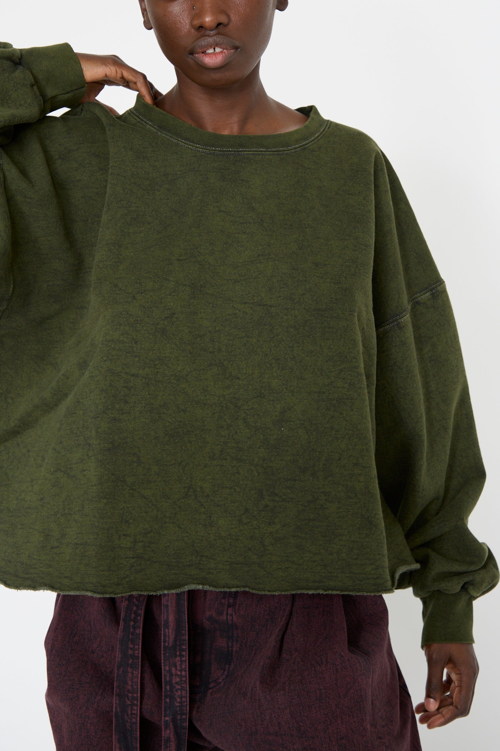 A woman is standing against a plain background and adjusting her collar, wearing the Fonder Sweatshirt in Olive from Rachel Comey and maroon pants. 