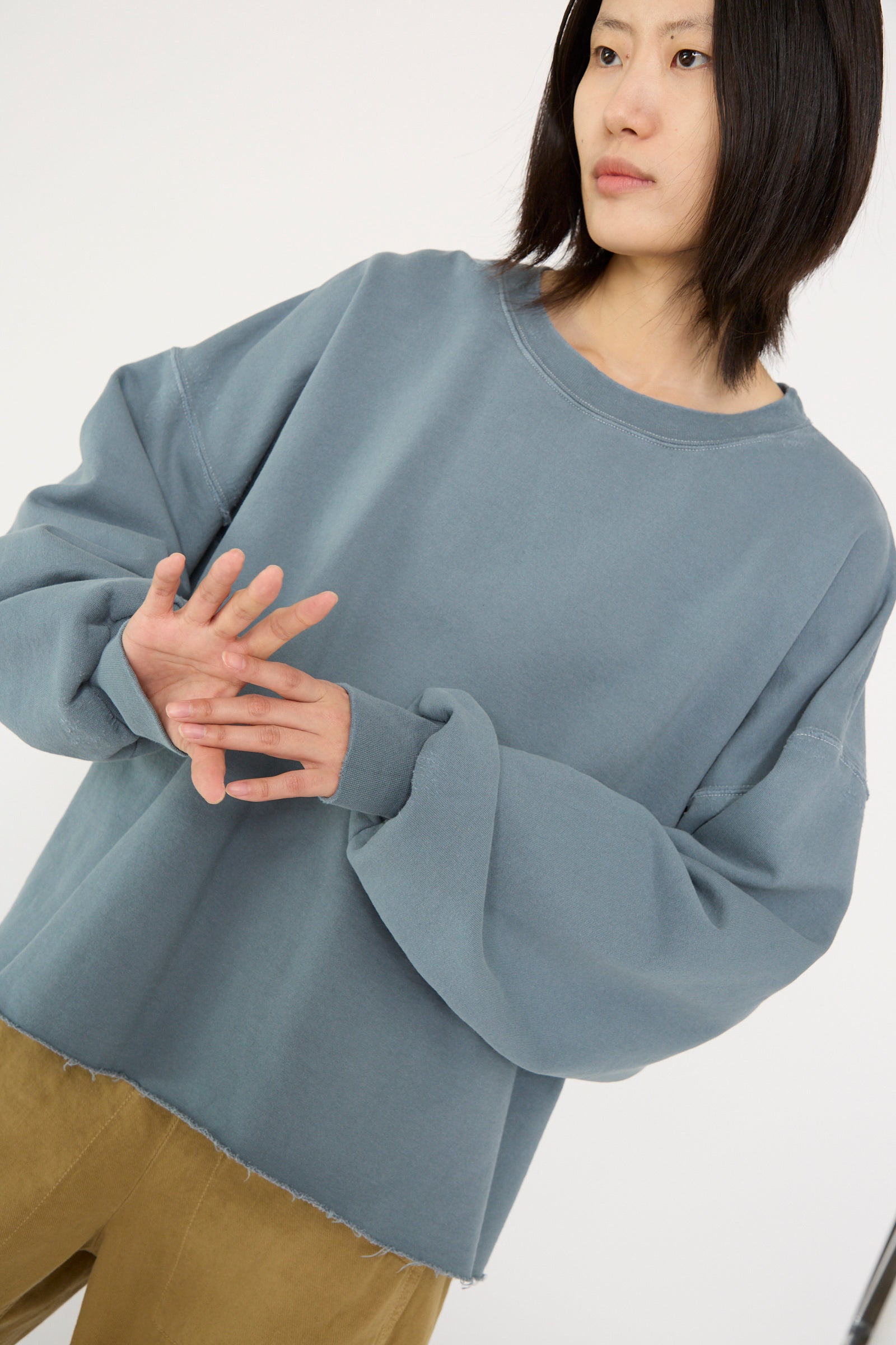 The model is wearing a loose, oversized fit Fonder Sweatshirt in Petrol by Rachel Comey and brown pants, with hands slightly extended, looking to the side against a plain background.