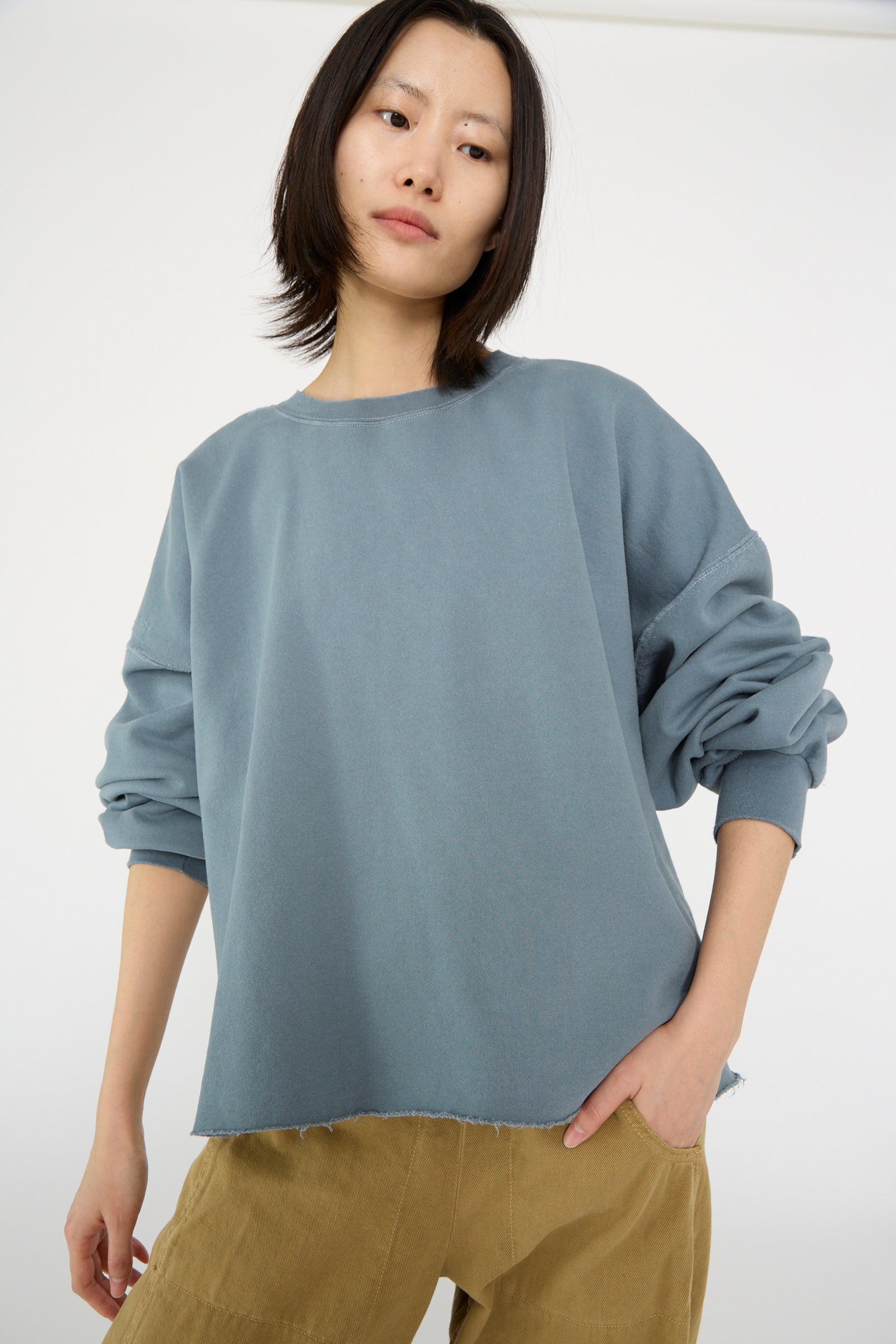 A person wearing the Rachel Comey Fonder Sweatshirt in Petrol and beige pants stands with one hand in their pocket against a plain white background.