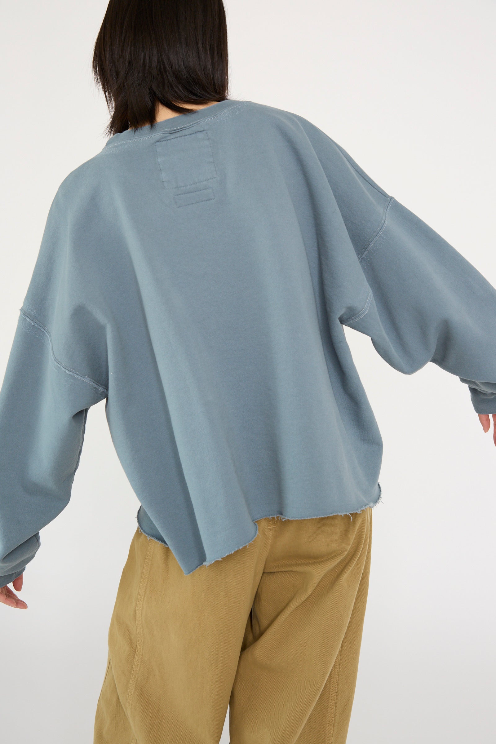 A person is wearing an oversized Fonder Sweatshirt in Petrol by Rachel Comey with brown pants, facing away and slightly extending their arms.