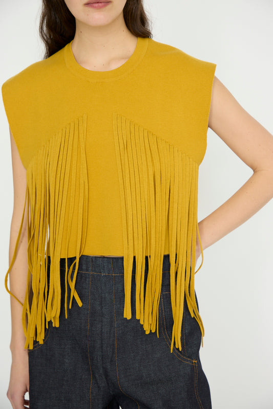 A person models the Fringed Briones Top in Mustard by Rachel Comey, its fringe draping elegantly over dark jeans, with arms relaxed at their sides against a plain background.