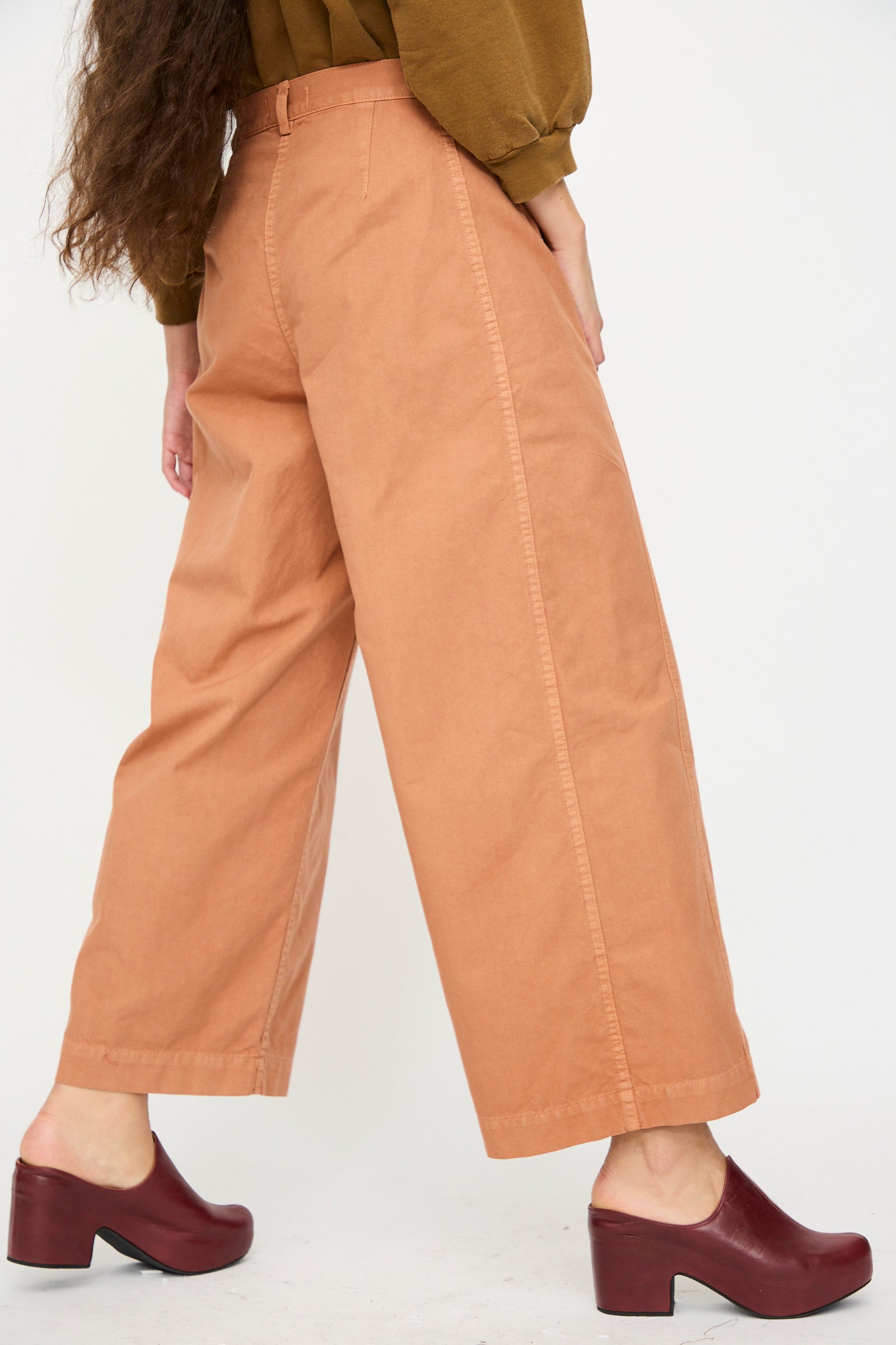 A person wears the Rachel Comey Garra Pant in Copper with a brown long-sleeve top and dark red clogs, standing against a plain white background.